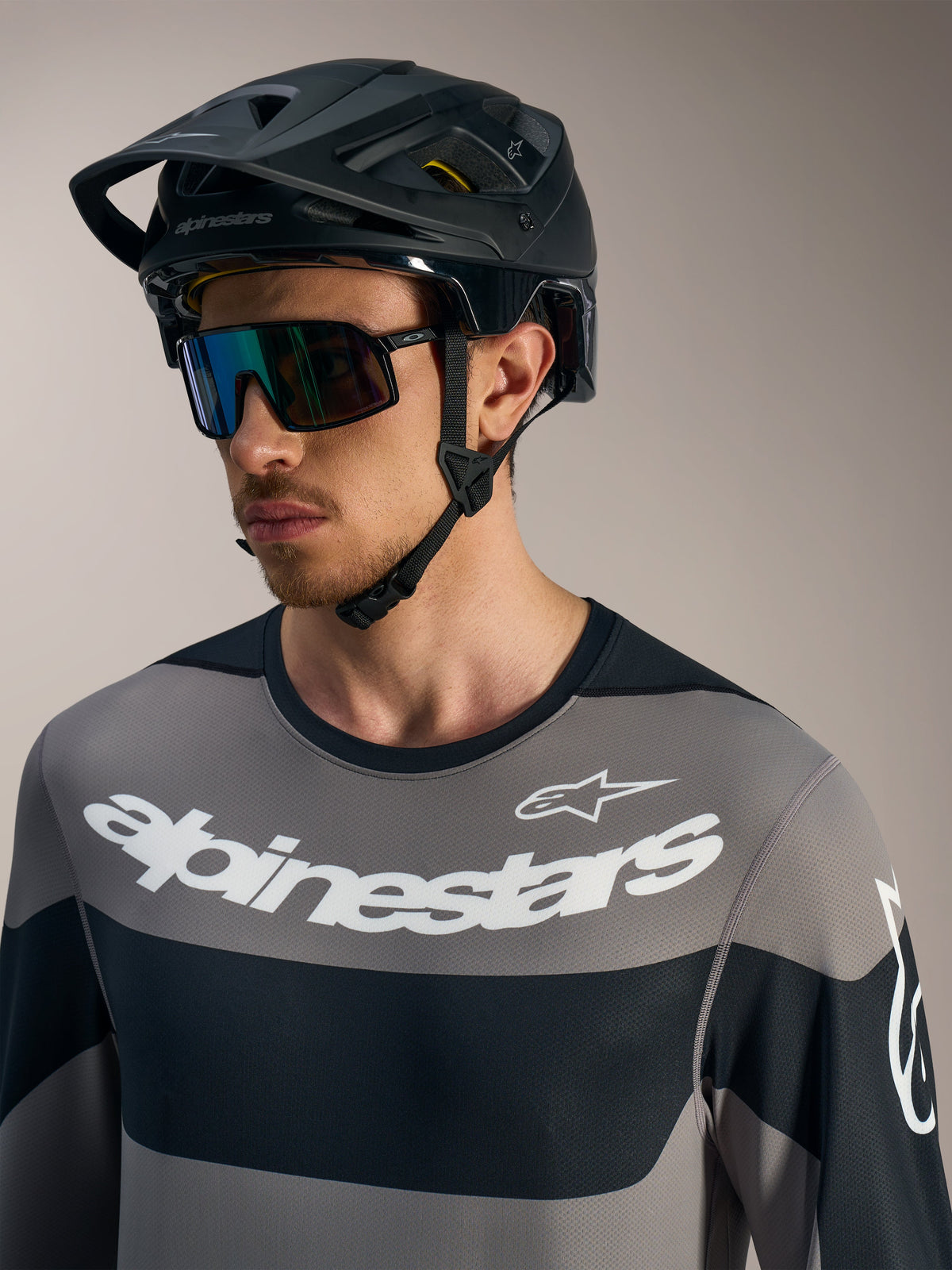 Camisola Alpinestars A-Dura Race cinzenta com logótipos brancos destacados, capacete de BTT preto e óculos de performance.