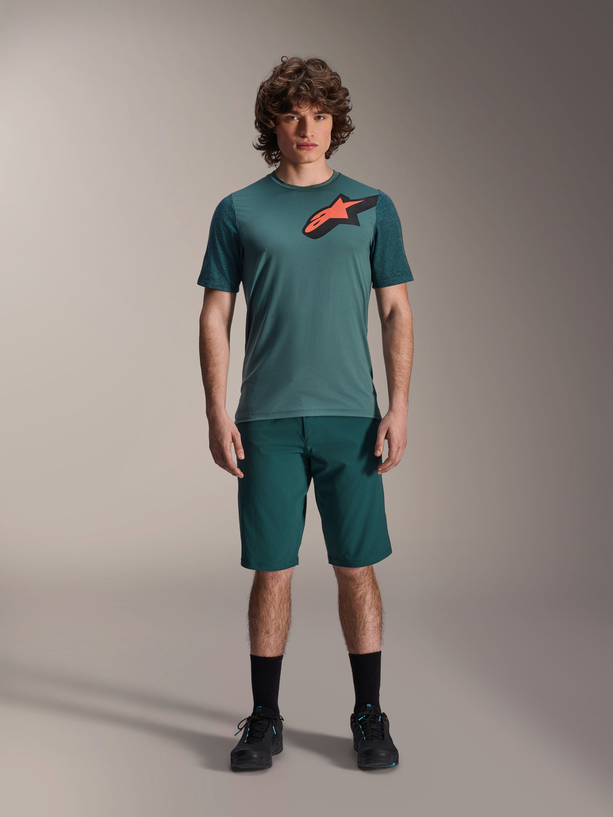 Alpinestars A-Dura Shorts in Dark Pine, men's mountain bike shorts, shown on a model with a matching teal short-sleeve jersey featuring a red and black star logo, black socks and cycling shoes, casual sporty mountain biking apparel