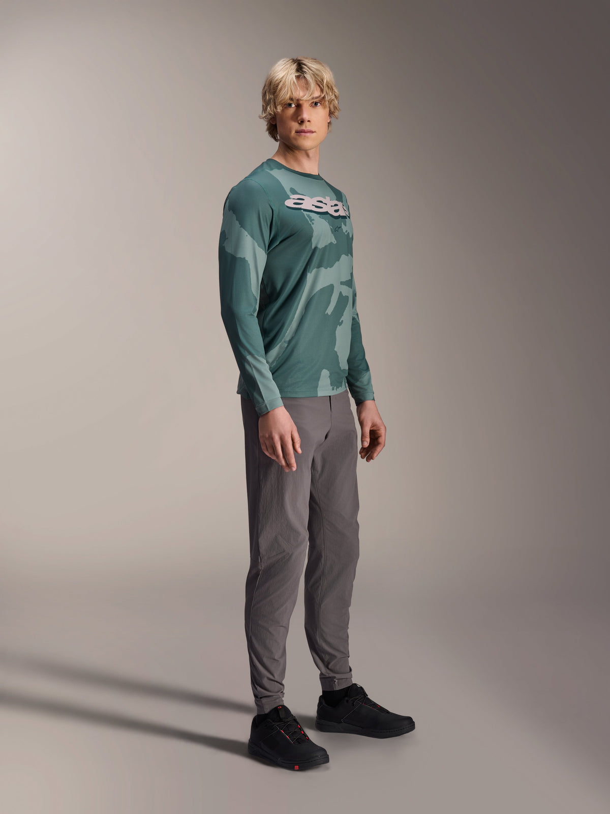 Camisola de Manga Comprida Alpinestars A-Dura Hollow Camo, Silver Pine Camo, vestida por um modelo com calças cinzentas e sapatos pretos, com um padrão de camuflagem verde e um logótipo ASTARS branco em destaque no peito, vestuário de BTT de alta performance