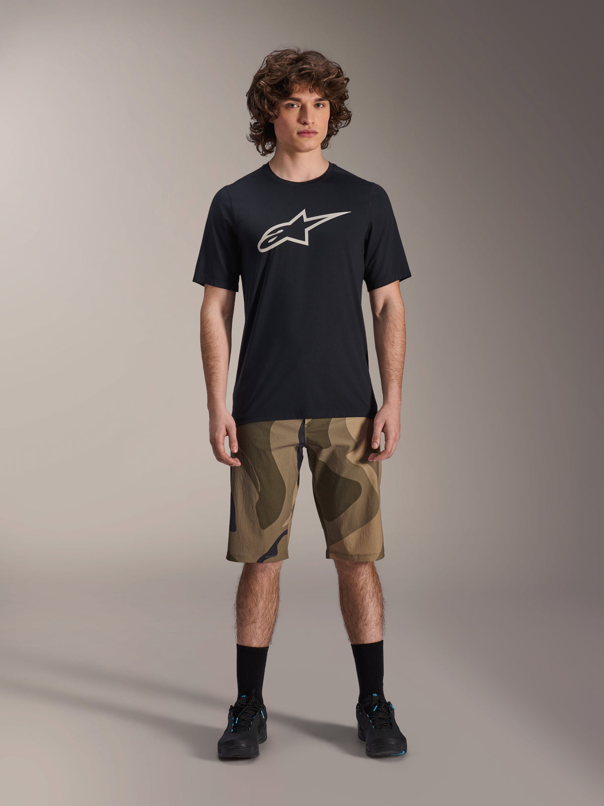 Camisola de Manga Curta Alpinestars A-Dura Astar, preta com logótipo Astars branco grande no peito, vestida por um modelo com calções de BTT camuflados e sapatilhas de ciclismo pretas, vestuário casual de BTT