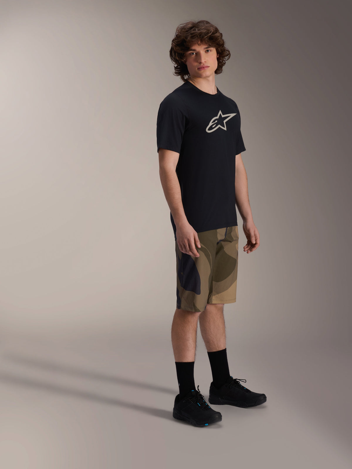 Camisola de Manga Curta Alpinestars A-Dura Astar, preta com logótipo branco no peito, usada com calções camuflados e sapatos de BTT pretos, equipamento casual para ciclismo de montanha