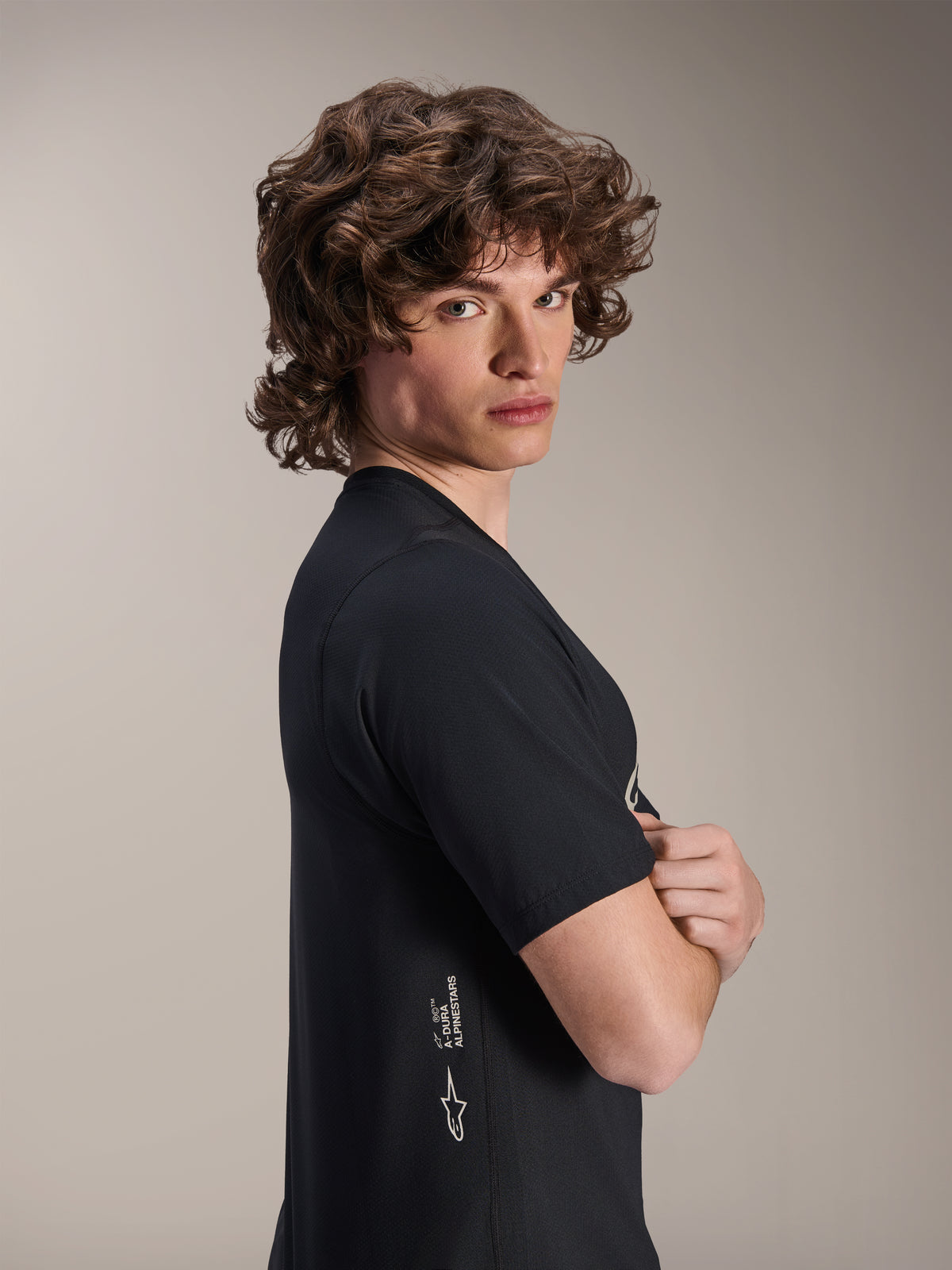 Alpinestars A-Dura Astar Jersey, camisola de BTT de manga curta, preta, modelo masculino posando de perfil com os braços cruzados, com logótipo Alpinestars a branco na parte lateral inferior, design atlético casual para ciclismo de montanha.