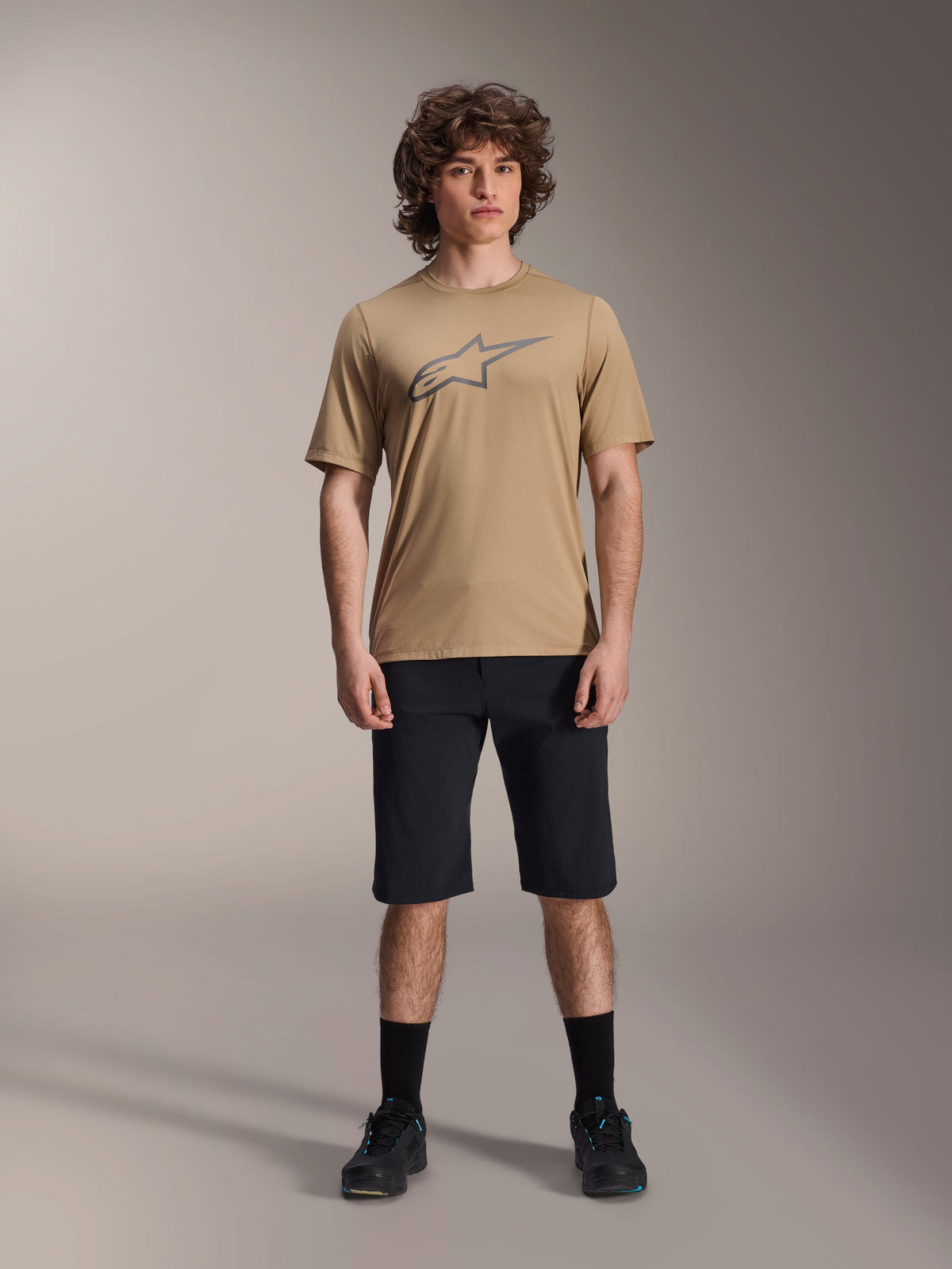 Alpinestars A-Dura Astar Short Sleeve Jersey, castanho claro, usada com calções pretos e sapatos pretos, com um logótipo Astar delineado de grandes dimensões no peito, tecido jacquard de alta performance para trilhos de mountain bike