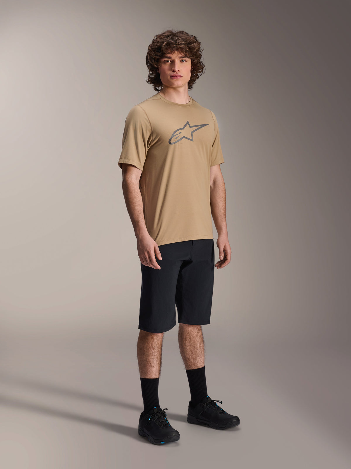 Camisola Alpinestars A-Dura Astar de Manga Curta, Castanho Claro, com um logótipo Astar grande em cinzento escuro no peito, vestida por um modelo com calções de MTB pretos, meias pretas e sapatos pretos, vestuário casual de alta performance para mountain bike em ambiente de estúdio