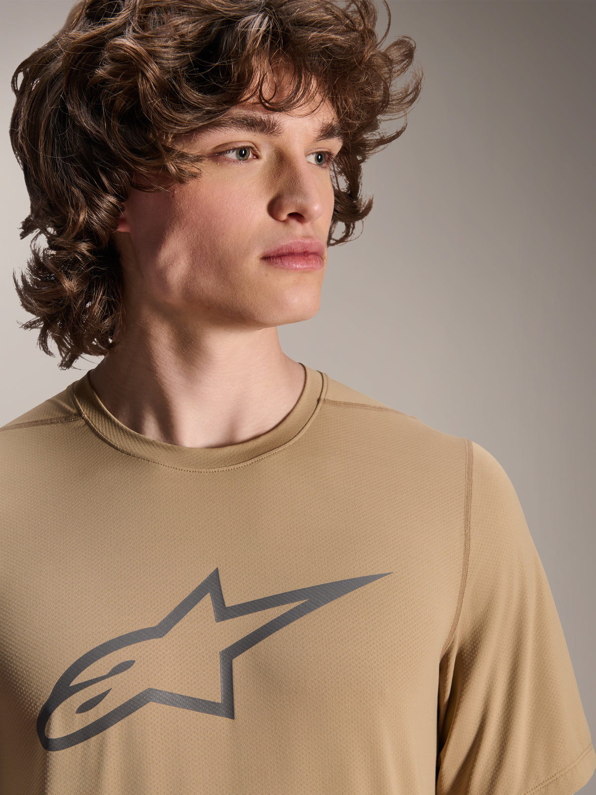 Modelo a usar a Camisola de Manga Curta Alpinestars A-Dura Astar, camisola de BTT castanho claro, com logótipo de estrela cinzento no peito, tecido jacquard que afasta a humidade, estilo de ciclismo casual