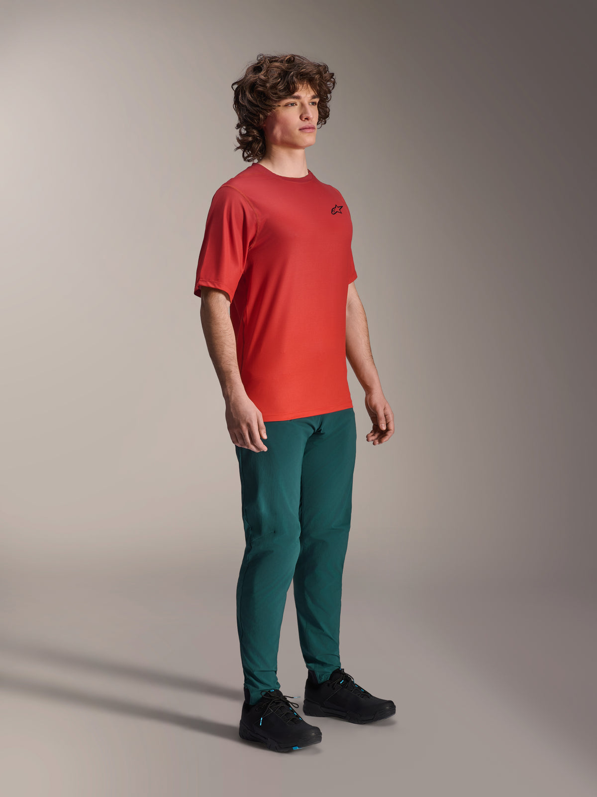 Modelo a usar calças Alpinestars A-Dura em Dark Pine, combinadas com uma camisola de manga curta A-Dura Rise vermelha e sapatos de BTT pretos, vestuário desportivo de mountain bike em ambiente de estúdio.