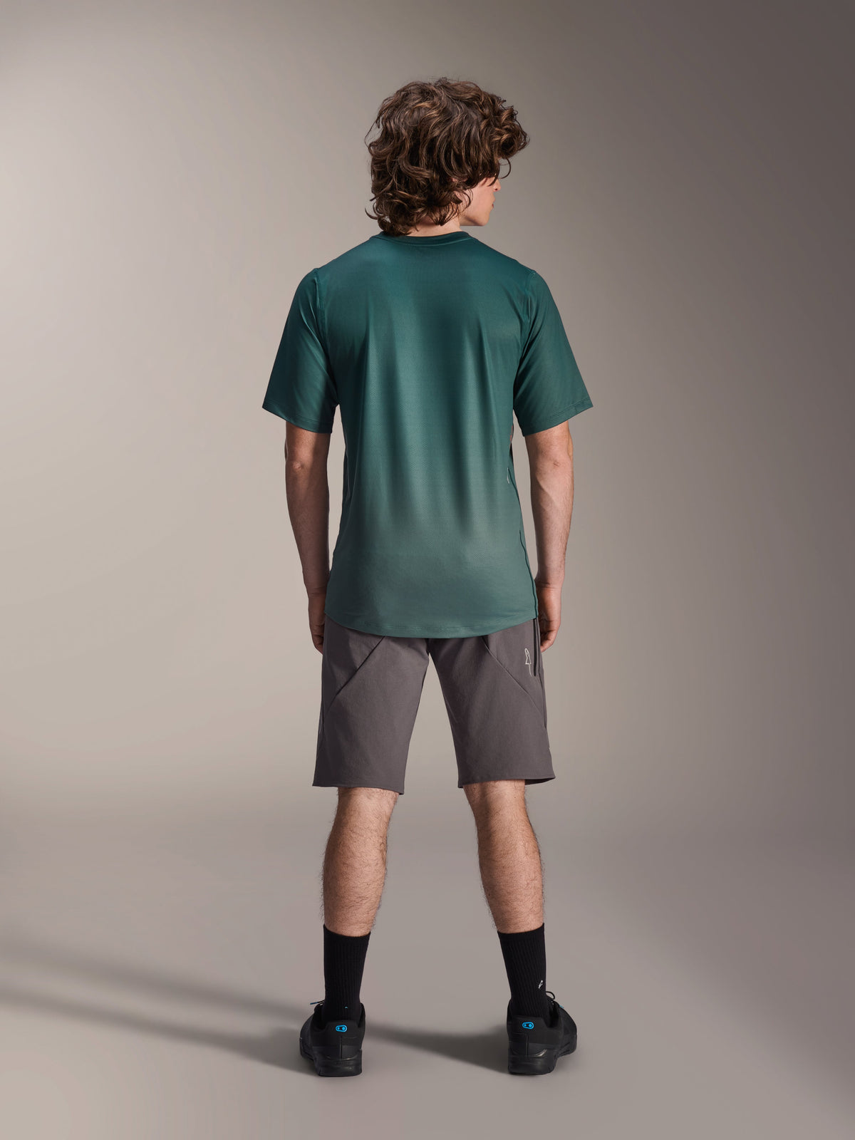 Camisola Alpinestars A-Dura Rise - Manga Curta, Verde Silver Pine, vista traseira de um modelo masculino a usar a camisola com calções cinzentos e meias pretas, apresentando uma cauda mais longa e ajuste regular para ciclismo de montanha