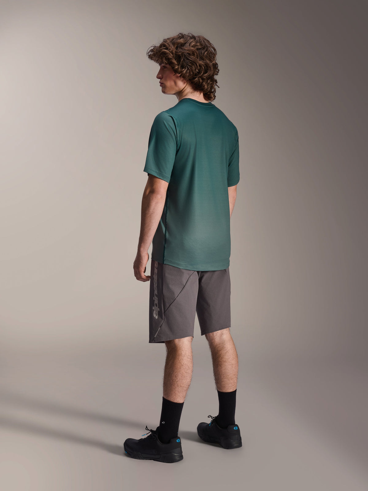 Alpinestars A-Dura Rise Jersey, camisola de BTT de manga curta, verde Silver Pine, usada com calções cinzentos e sapatos pretos, vista traseira mostrando a parte de trás mais comprida e o ajuste regular, tecido leve com absorção de humidade para trilhos