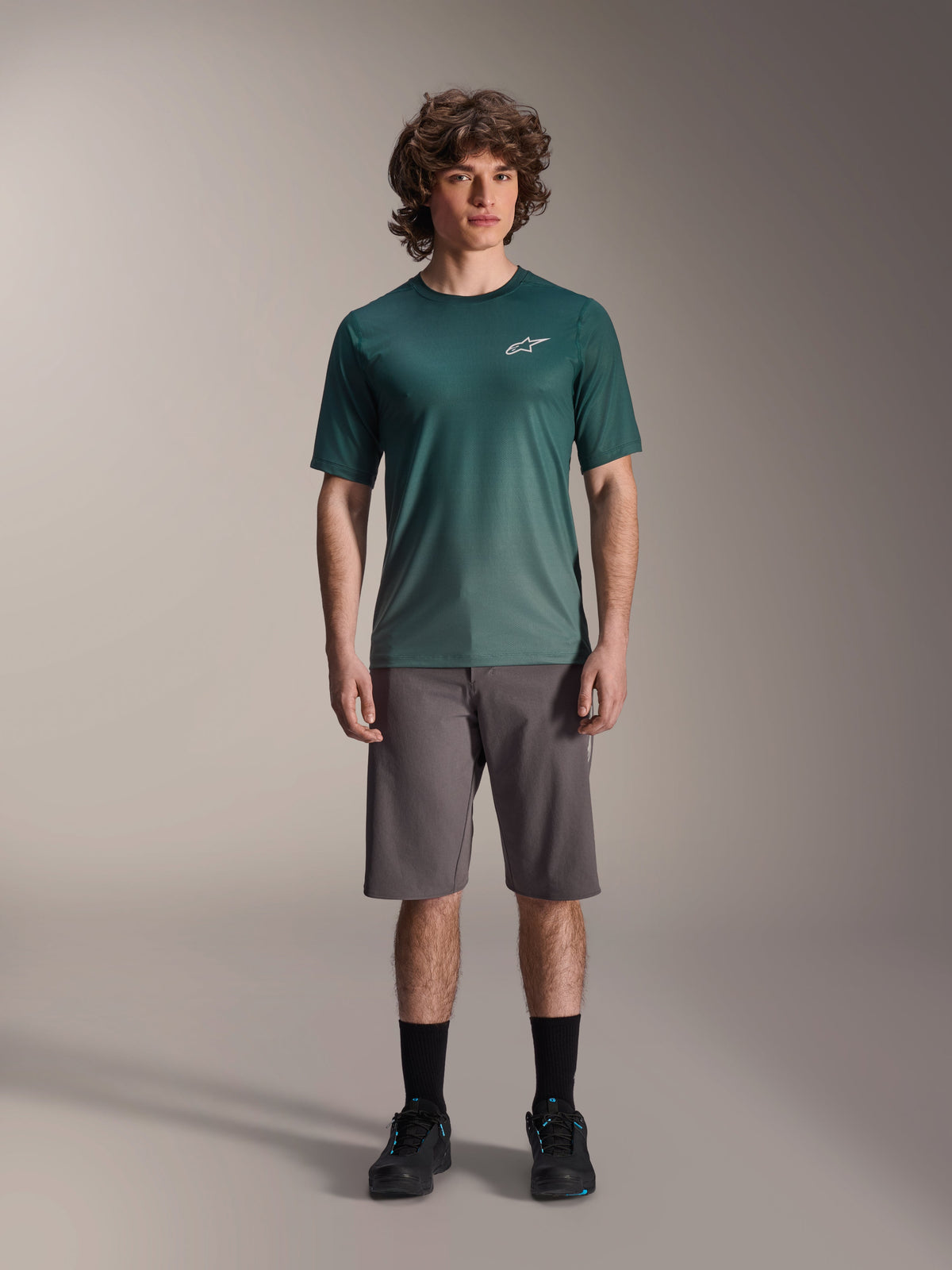Modelo masculino a usar a Camisola de Manga Curta Alpinestars A-Dura Rise em Silver Pine, camisola de ciclismo verde com pequeno logótipo branco no peito, combinada com calções de BTT cinzentos e calçado preto, equipamento casual para trilhos de BTT