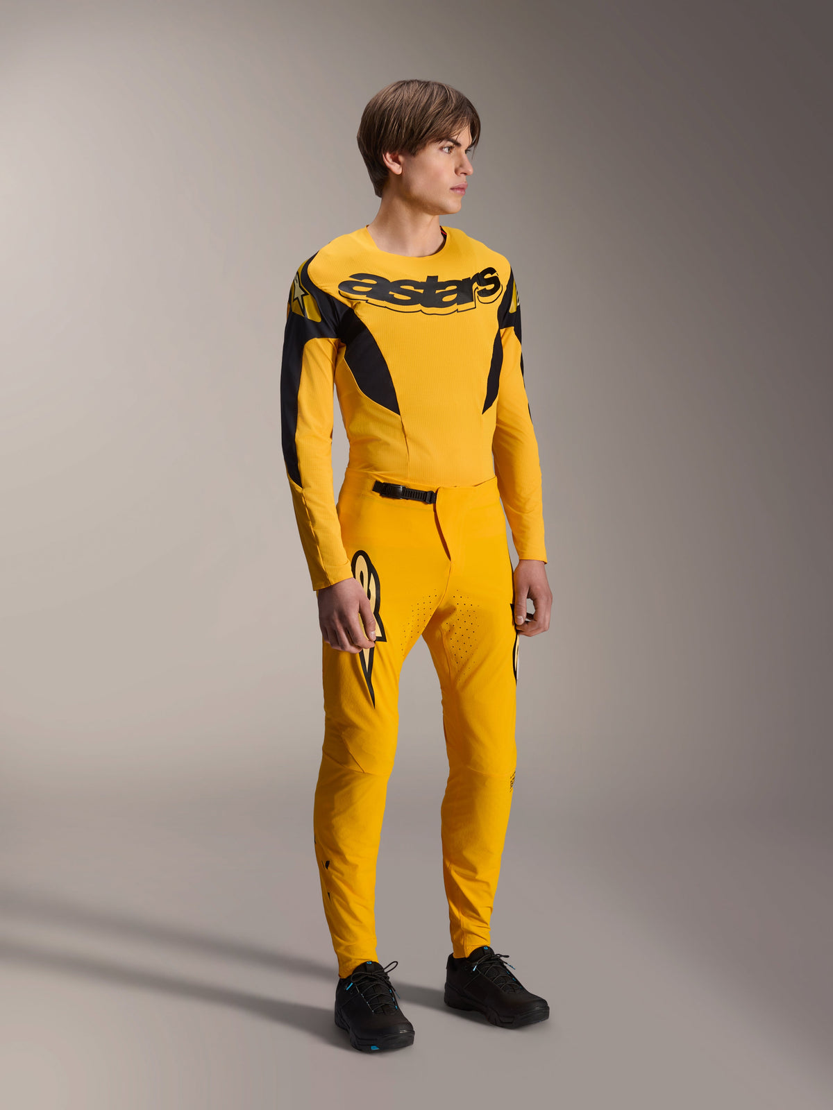 Modelo a usar a camisola Alpinestars Limited Edition A-Supra Melt Gold, camisola de MTB de manga comprida em amarelo escuro e preto, combinada com calças amarelas a condizer e sapatos pretos, equipamento técnico de ciclismo todo-o-terreno com gráficos inspirados na competição