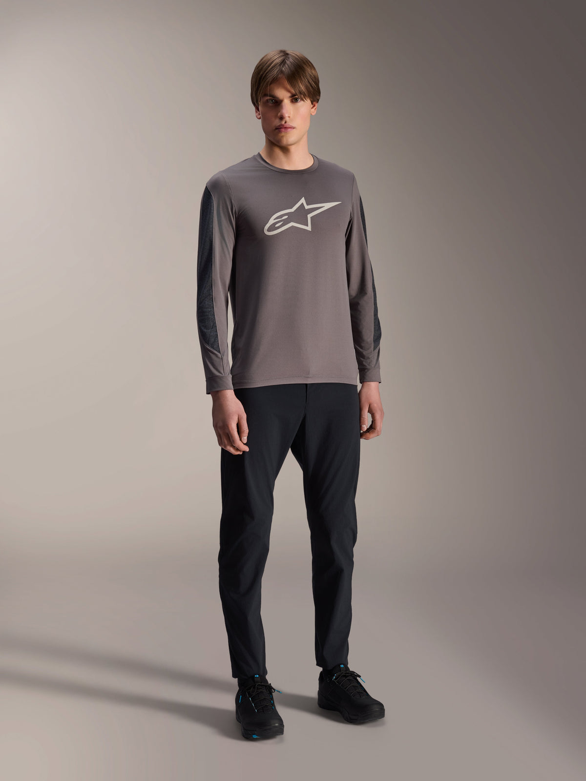 Alpinestars Camisola A-Dura Insert Astar, camisola de BTT de manga comprida, cinzento escuro, com grande logótipo branco no peito, usada com calças técnicas pretas e sapatos pretos, vestuário de ciclismo todo-o-terreno
