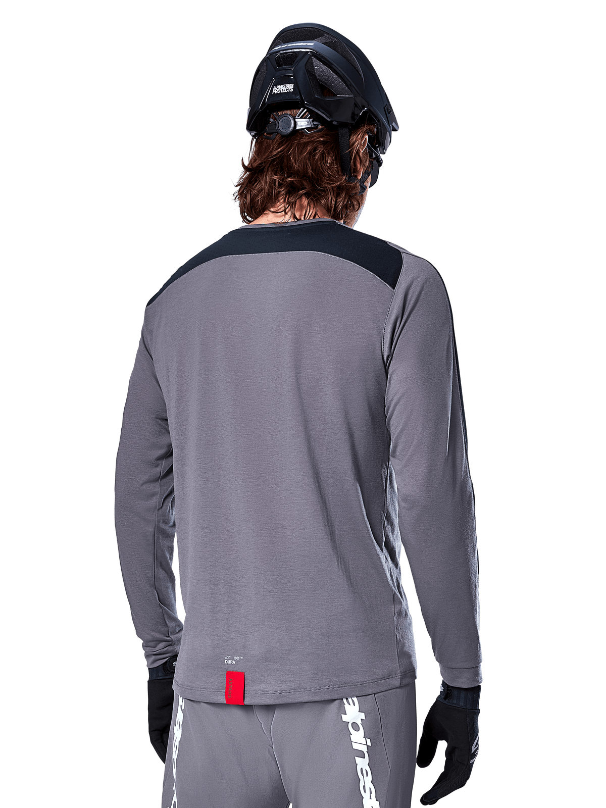 Camisola de Manga Comprida Alpinestars A-Dura Dri Astar, camisola de MTB cinza escuro vista de trás, com um painel preto nos ombros e uma pequena etiqueta vermelha da marca, usada com um capacete de mountain bike preto e luvas
