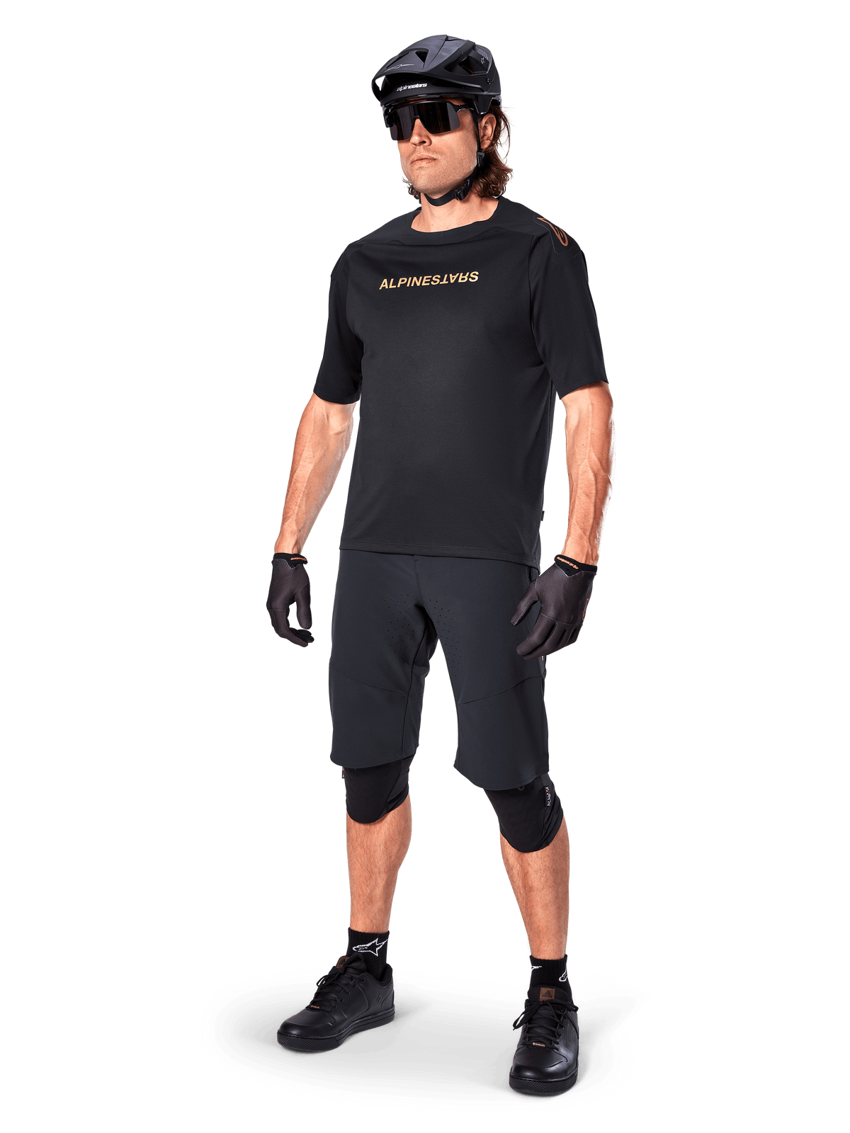 Alpinestars A-Aria Polartec Switch Short Sleeve Jersey, camisola de manga curta preta, com logótipo dourado espelhado no peito, usada por um ciclista com calções de ciclismo pretos, capacete e joelheiras, equipamento técnico de MTB para desempenho fora de estrada