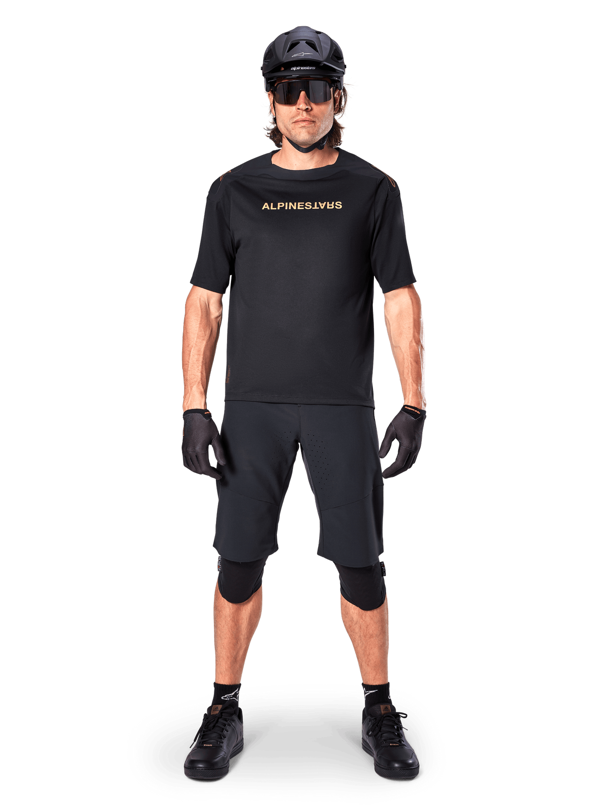 Alpinestars A-Aria Polartec Switch Jersey, camisola de BTT de manga curta, preta com letras beges, usada com equipamento de BTT preto coordenado incluindo capacete, calções e joelheiras, vestuário de ciclismo de alta performance para todo-o-terreno