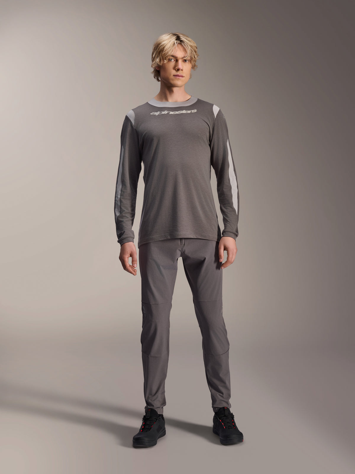 Camisola de Manga Comprida Alpinestars A-Dura Dri Wool, cinzento escuro, usada por modelo com calças cinzentas a condizer e sapatos pretos, logótipo Alpinestars branco no peito, painéis de malha cinzento claro nos ombros e mangas, equipamento técnico de mountain bike