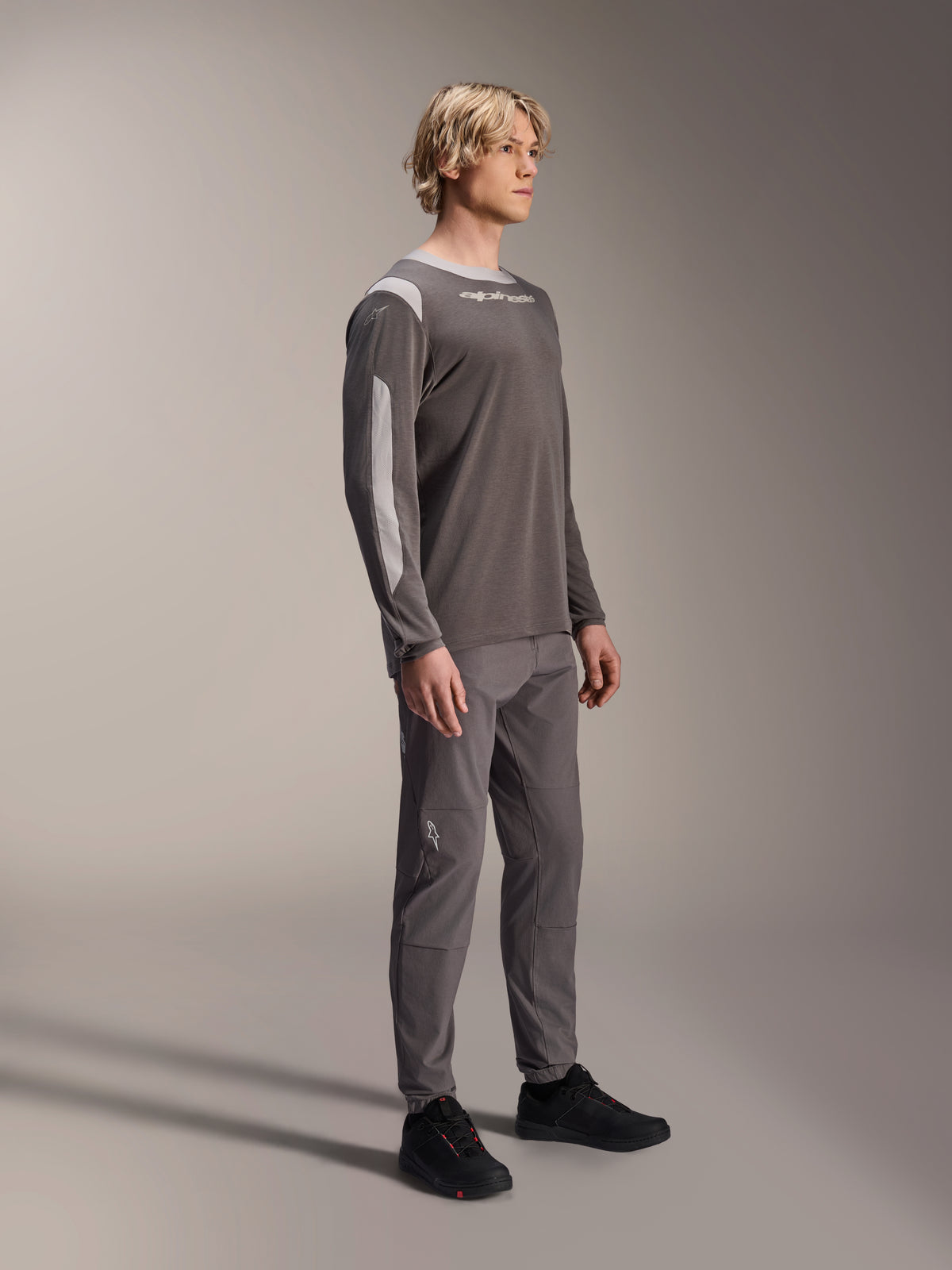 Modelo masculino a usar a camisola Alpinestars A-Dura Dri Wool de manga comprida em cinzento escuro, combinada com calças cinzentas a condizer e sapatos pretos, com painéis de malha cinzento claro nos ombros e mangas, equipamento técnico de BTT para ciclismo fora de estrada