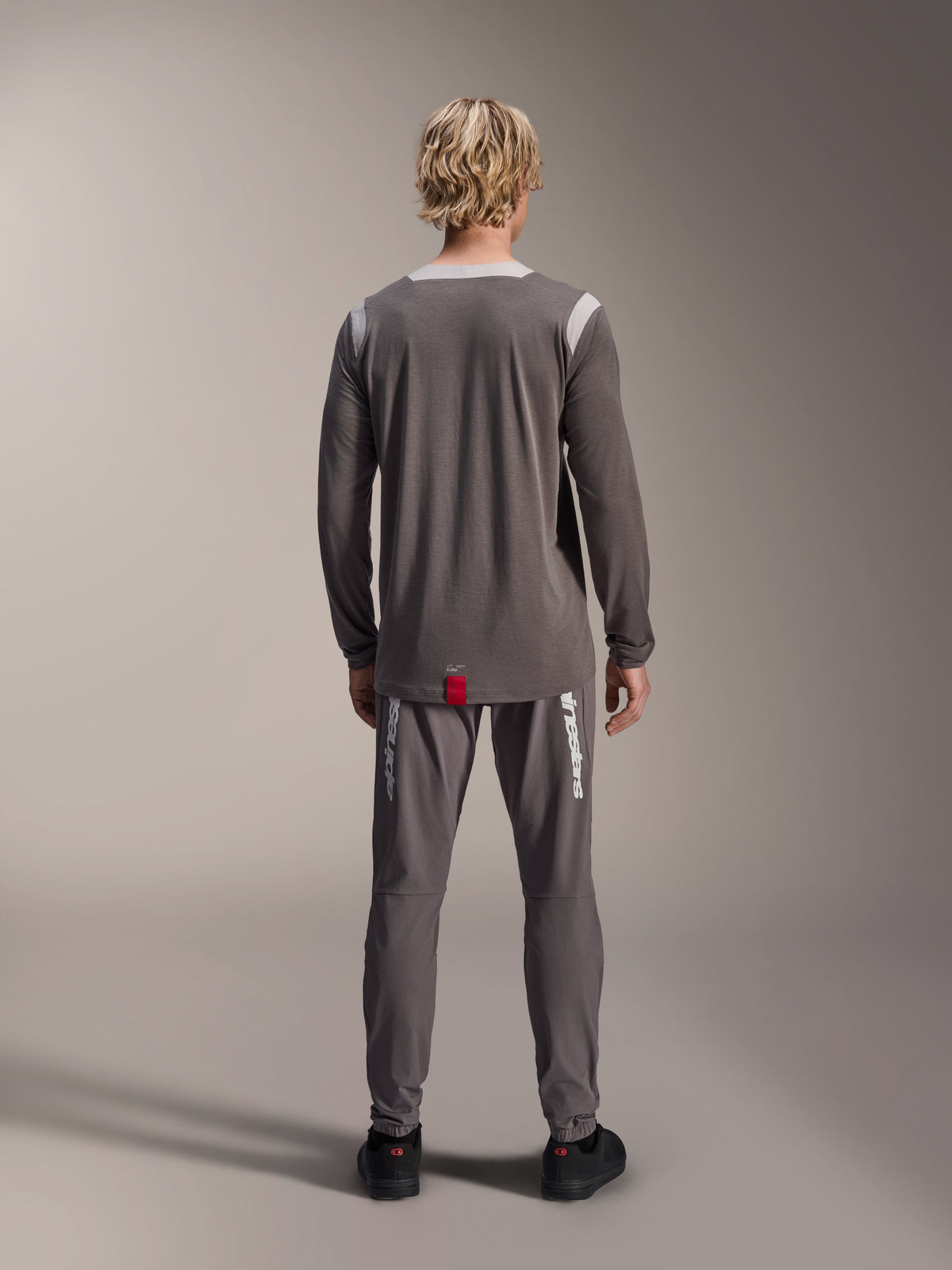 Alpinestars Camisola A-Dura Dri Wool - Manga Comprida, cinzento escuro, vista traseira num modelo, com painéis de malha cinzento claro nos ombros e gola, cauda mais longa com uma pequena etiqueta vermelha da marca, usada com calças de ciclismo cinzentas a condizer.