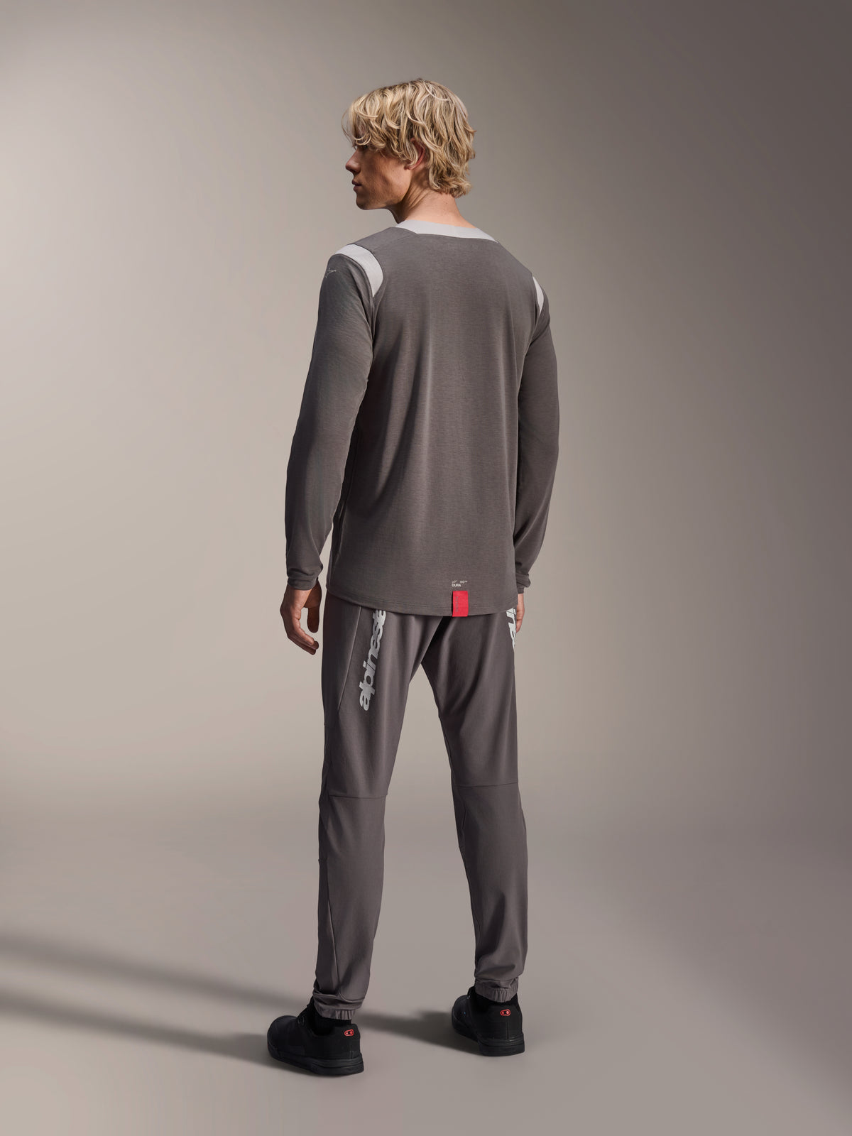 Modelo masculino a usar camisola de manga comprida Alpinestars A-Dura Dri Wool, cinzento escuro com painéis de malha cinzento claro nos ombros, combinada com calças de ciclismo cinzento escuro a condizer, vista de costas a mostrar a bainha traseira alongada, vestuário de BTT