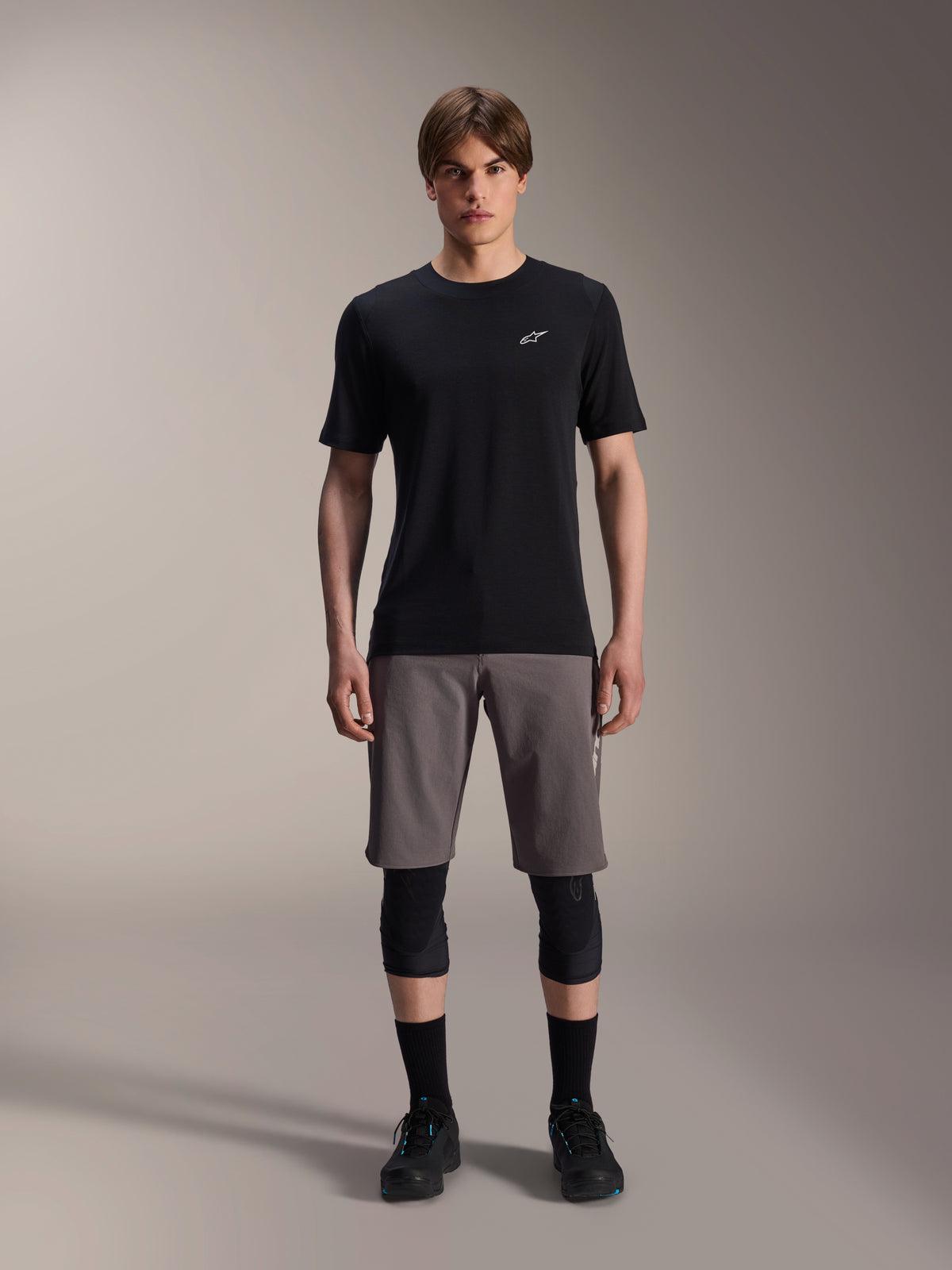 Male model wearing Alpinestars A-Dura Elite Shorts in Dark Gray and A-Dura Dri-Wool Short Sleeve Jersey in Black, paired with black knee guards and cycling shoes, technical mountain bike apparel for off-road performance.