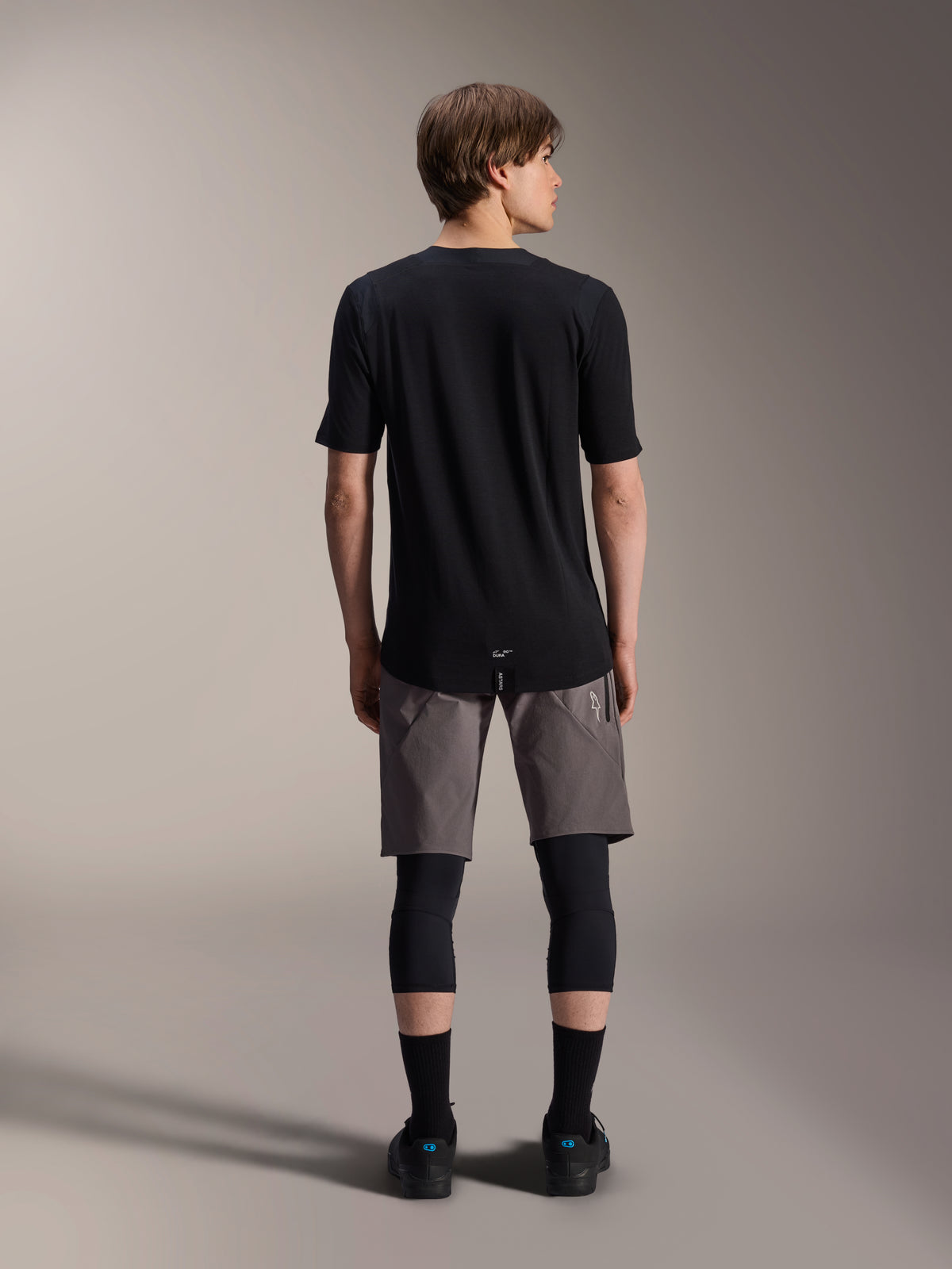 Alpinestars A-Dura Dri Wool Short Sleeve Jersey, Black, back view worn by model with grey MTB shorts and black knee guards, featuring a longer drop tail and athletic fit for off-road cycling