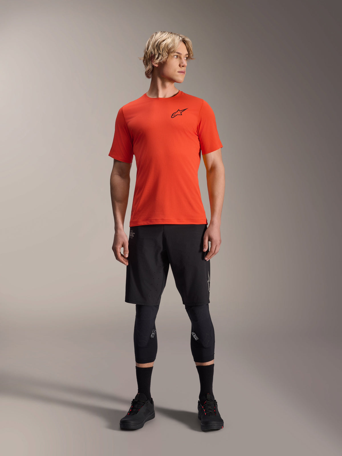 Alpinestars A-Aria Astar Short Sleeve Jersey, Spicy Orange, usada por modelo com calções de BTT pretos e joelheiras, vestuário de ciclismo de alta performance leve e respirável para BTT
