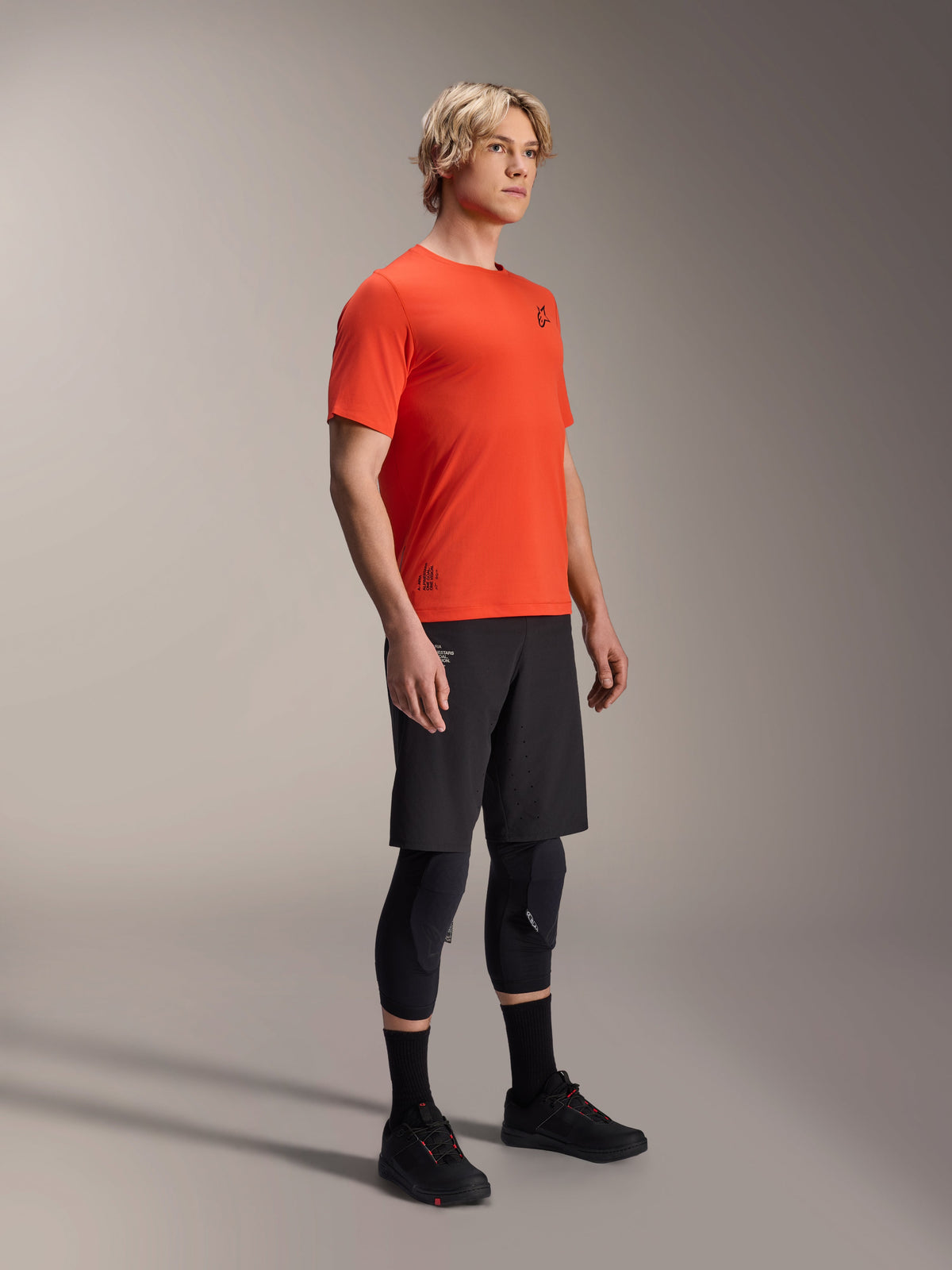 Camisola de Manga Curta Alpinestars A-Aria Astar em Spicy Orange, vestida por um modelo com calções de BTT pretos e proteções de joelhos, vestuário de BTT leve e respirável, laranja vibrante com logótipos pretos minimalistas