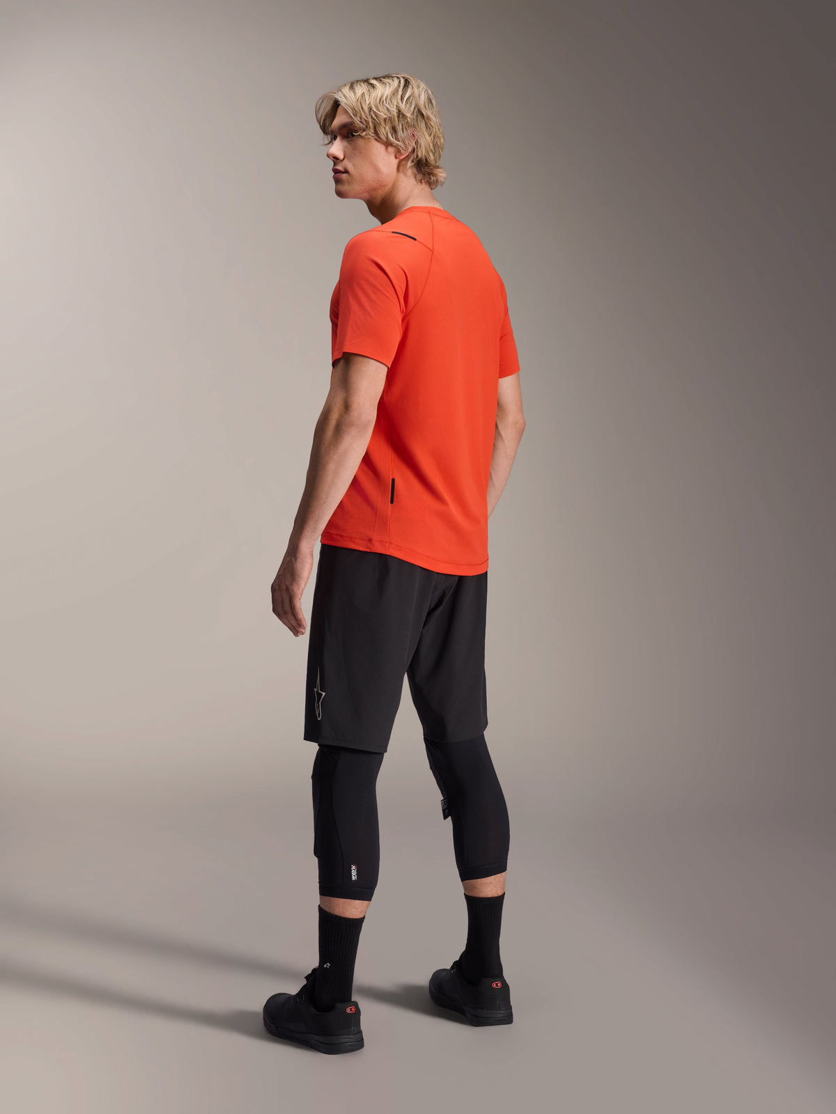 Camisola de Manga Curta Alpinestars A-Aria Astar, Spicy Orange, vista de três quartos de costas do modelo a olhar por cima do ombro, combinada com calções de BTT pretos e joelheiras, vestuário de performance para BTT