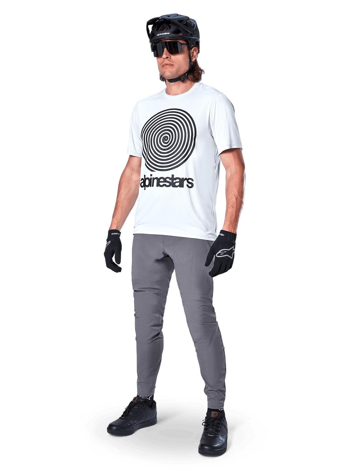 Alpinestars A-Dura Oscar Jersey - Manga Curta, cor White Dove com um grande gráfico espiral preto, vestida por um ciclista com calças de BTT cinzentas, capacete e luvas pretos, vestuário de ciclismo de alta performance para off-road