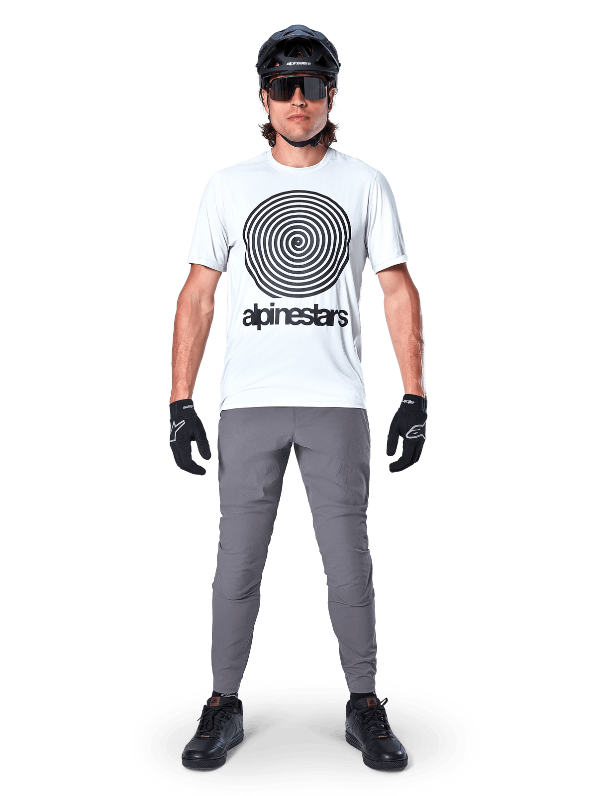 Ciclista de montanha a usar camisola de manga curta Alpinestars A-Dura Oscar, cor White Dove com grande logótipo em espiral preto, combinada com calças cinzentas, capacete preto, óculos de sol e luvas pretas, equipamento de alta performance para BTT