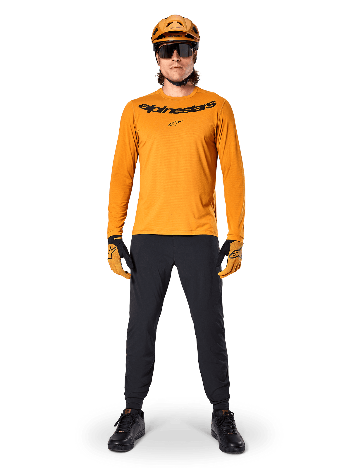 Alpinestars A-Dura Rocker Long Sleeve Jersey in Dark Gold, featuring a black screen-printed logo on the chest, worn with a matching gold MTB helmet, black sunglasses, and black pants, professional mountain biking gear