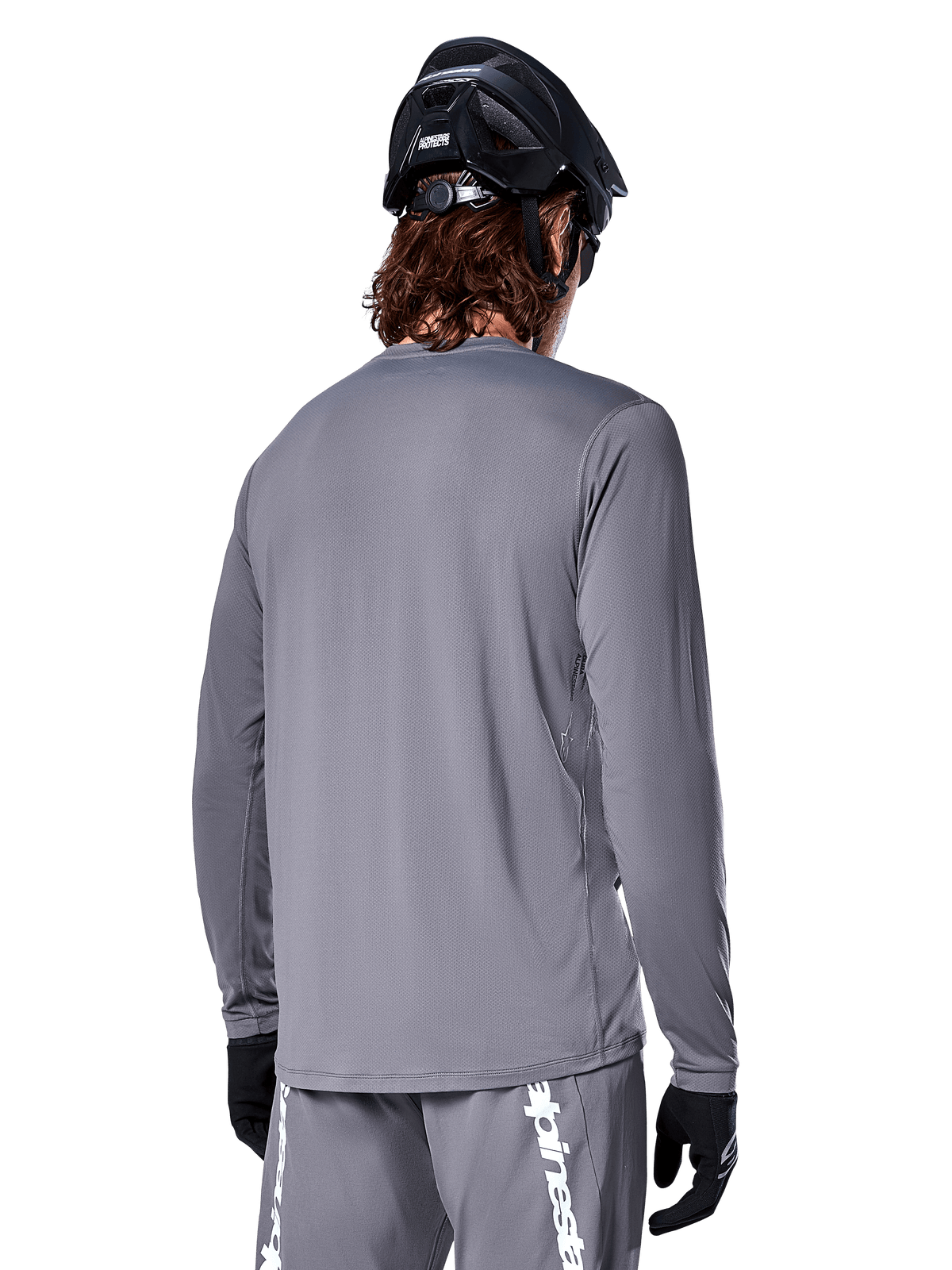 Alpinestars A-Dura Rocker Long Sleeve Jersey em Cinza Escuro, vista traseira de ciclista de BTT, combinada com calções cinzentos a condizer com logótipo branco, capacete de BTT preto e luvas pretas, equipamento profissional de ciclismo todo-o-terreno