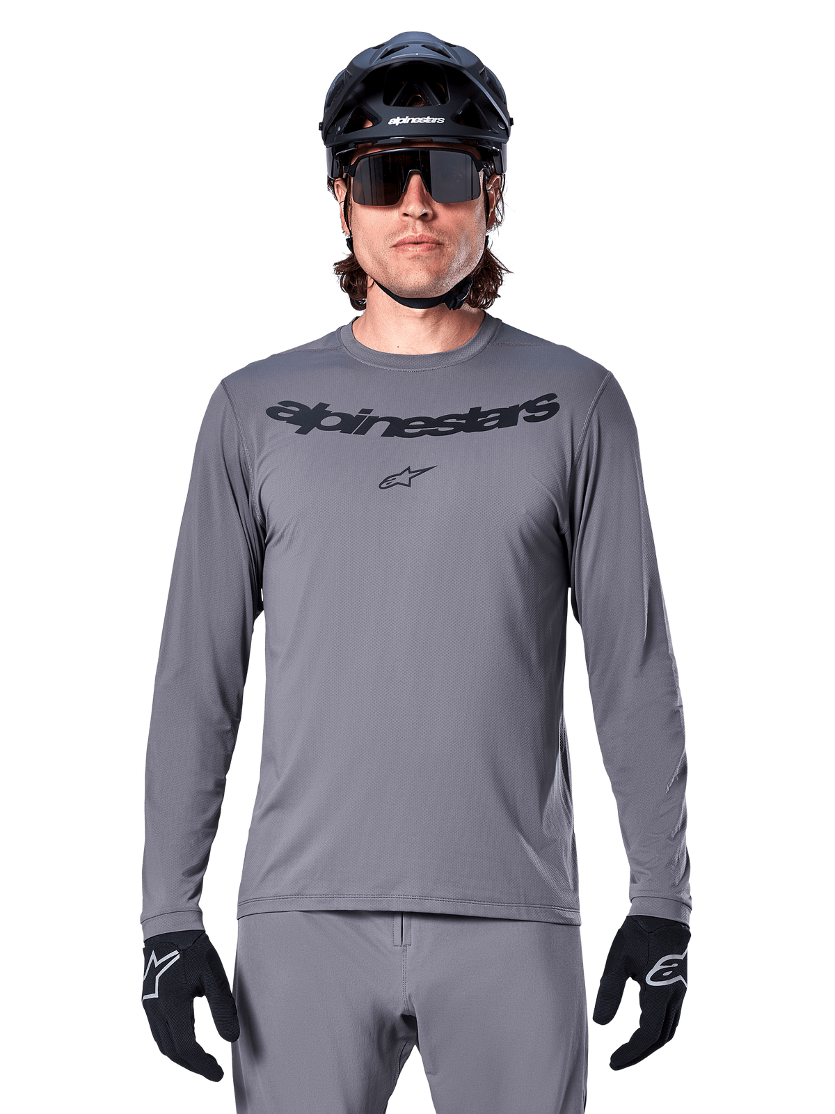 Ciclista masculino a usar a Camisola Alpinestars A-Dura Rocker de Manga Comprida, camisola de BTT cinzento-escuro com logótipo preto no peito, combinada com calças cinzentas, capacete e luvas pretos, desenhada para ciclismo fora de estrada
