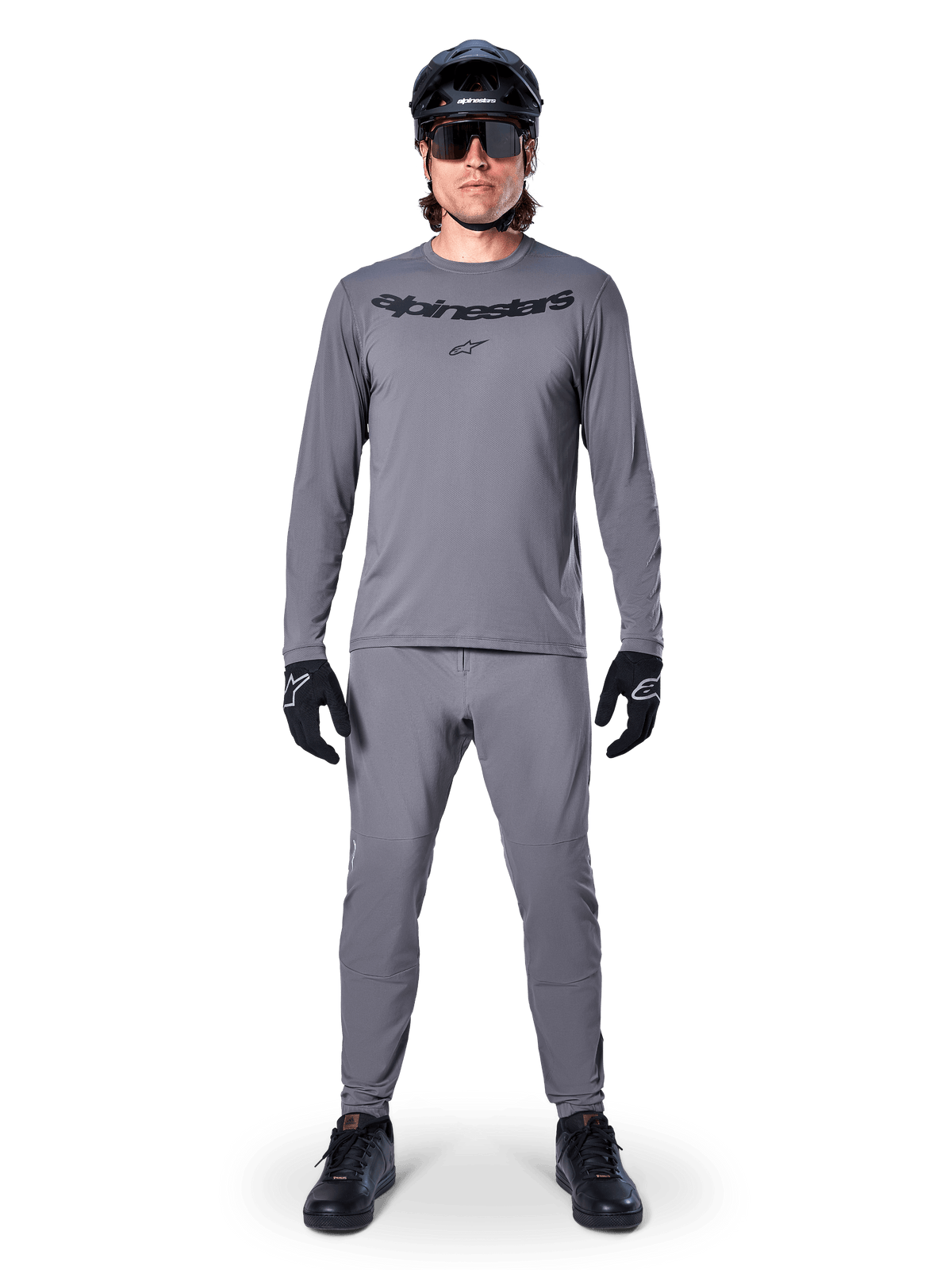 Ciclista com Camisola de Manga Comprida Alpinestars A-Dura Rocker em cinzento escuro, calças cinzentas a condizer, capacete de BTT preto, óculos de sol e luvas, vestuário profissional de BTT para trilhos fora de estrada