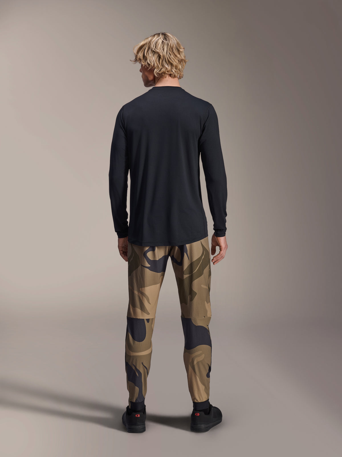 Alpinestars A-Dura Camo Pant, calças de Mountain Bike, padrão de camuflagem castanho, vista traseira, usadas com uma camisola preta de manga comprida e ténis pretos, calças têxteis de corte regular para ciclismo todo-o-terreno