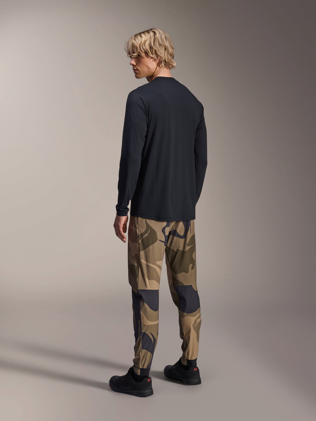 Alpinestars A-Dura Camo Pant, calças de MTB, camuflado castanho, usadas com uma camisola de manga comprida preta e sapatilhas pretas, vista traseira mostrando pernas afuniladas e punhos elásticos, vestuário de performance para ciclismo off-road