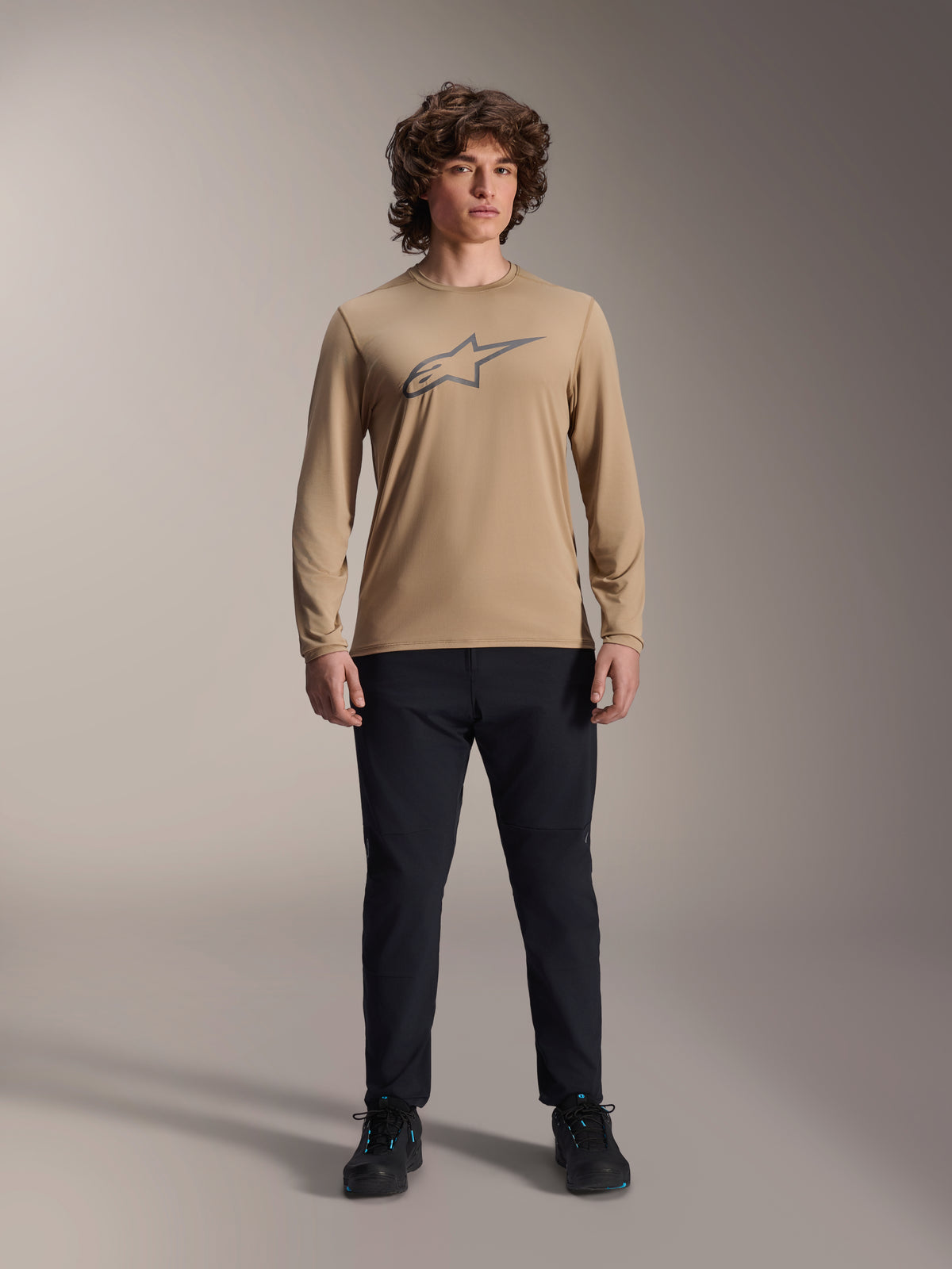 Camisola de Manga Comprida Alpinestars A-Dura Astar, castanho claro com logótipo Astars cinzento no peito, usada por um modelo com calças técnicas e sapatos pretos, equipamento casual de alta performance para mountain bike