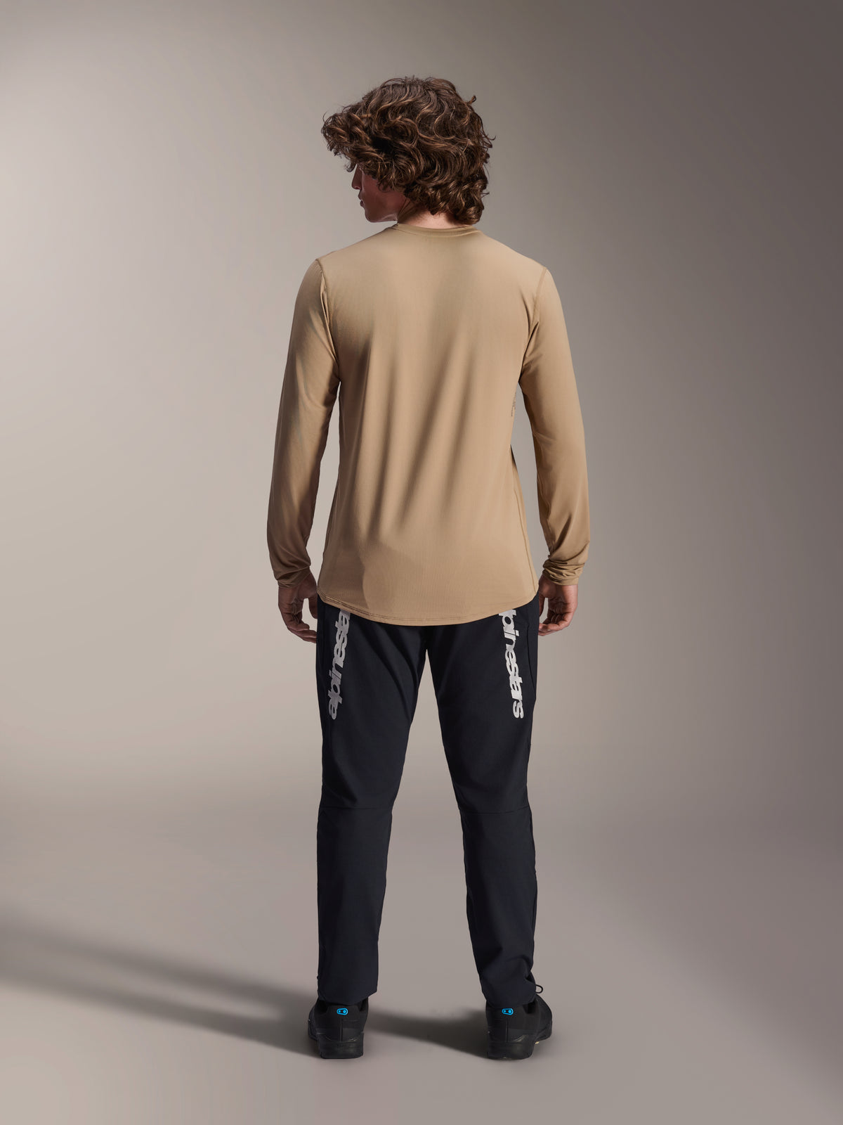 Camisola de Manga Comprida Alpinestars A-Dura Astar, castanho claro, vista traseira mostrando a cauda mais longa, usada com calças pretas com a marca Alpinestars, vestuário de alta performance para bicicleta de montanha