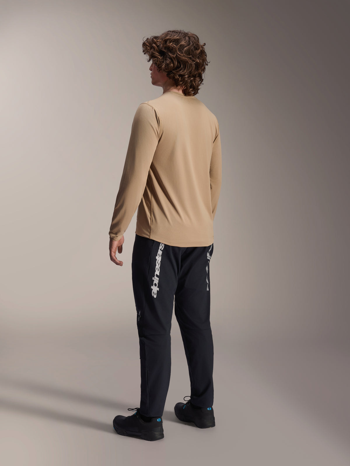 Camisola Alpinestars A-Dura Astar de Manga Comprida em Castanho Claro, vista traseira de um modelo masculino a exibir o design de cauda longa, combinada com calças e sapatos pretos, equipamento de performance casual para bicicleta de montanha.