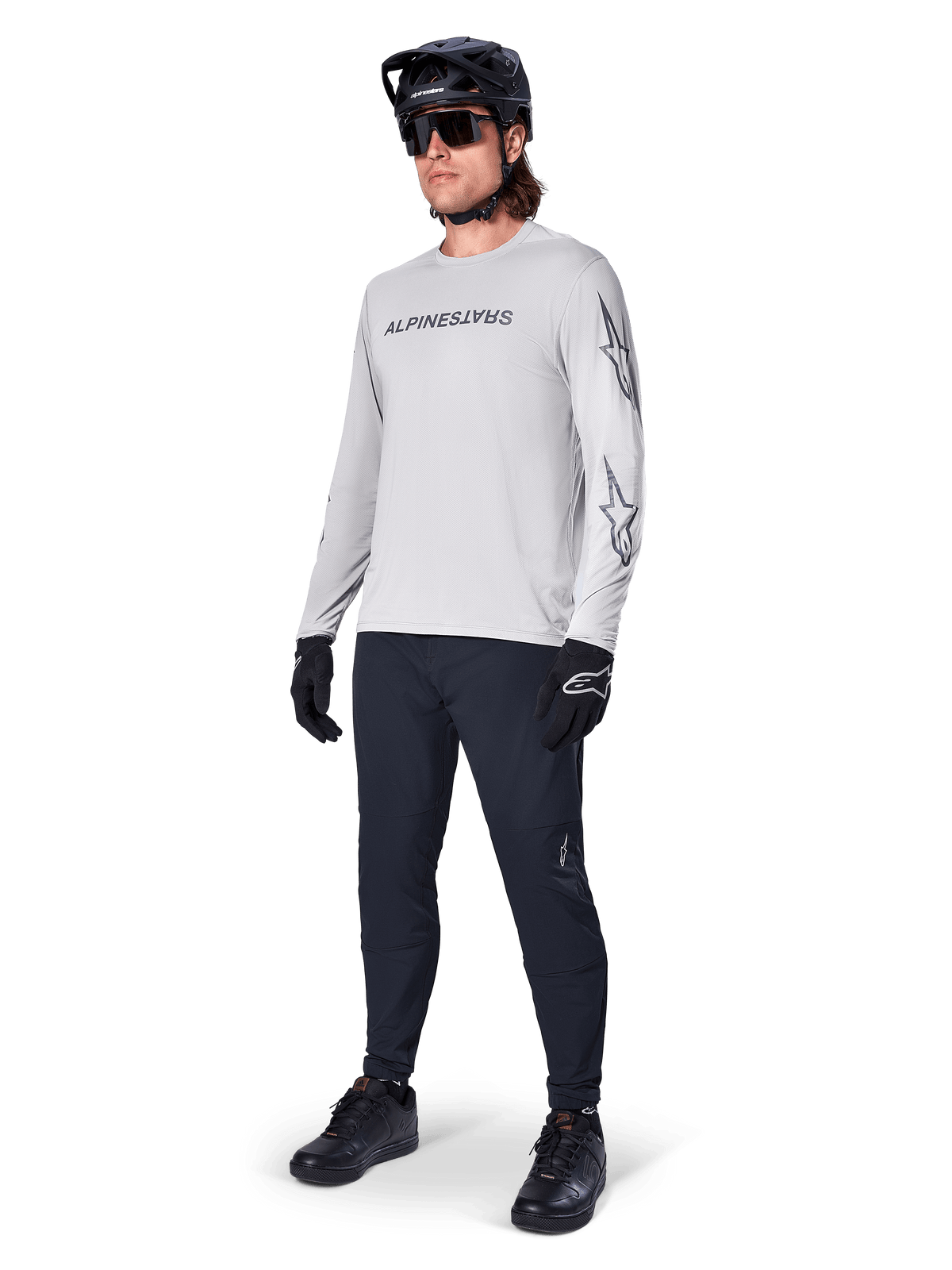 Camisola de Manga Comprida Alpinestars A-Dura Switch, cinzento claro, usada por ciclista masculino com calças de BTT pretas e capacete, com logótipo nominativo preto no peito e grafismos de estrela nas mangas, equipamento de ciclismo de alta performance para todo-o-terreno