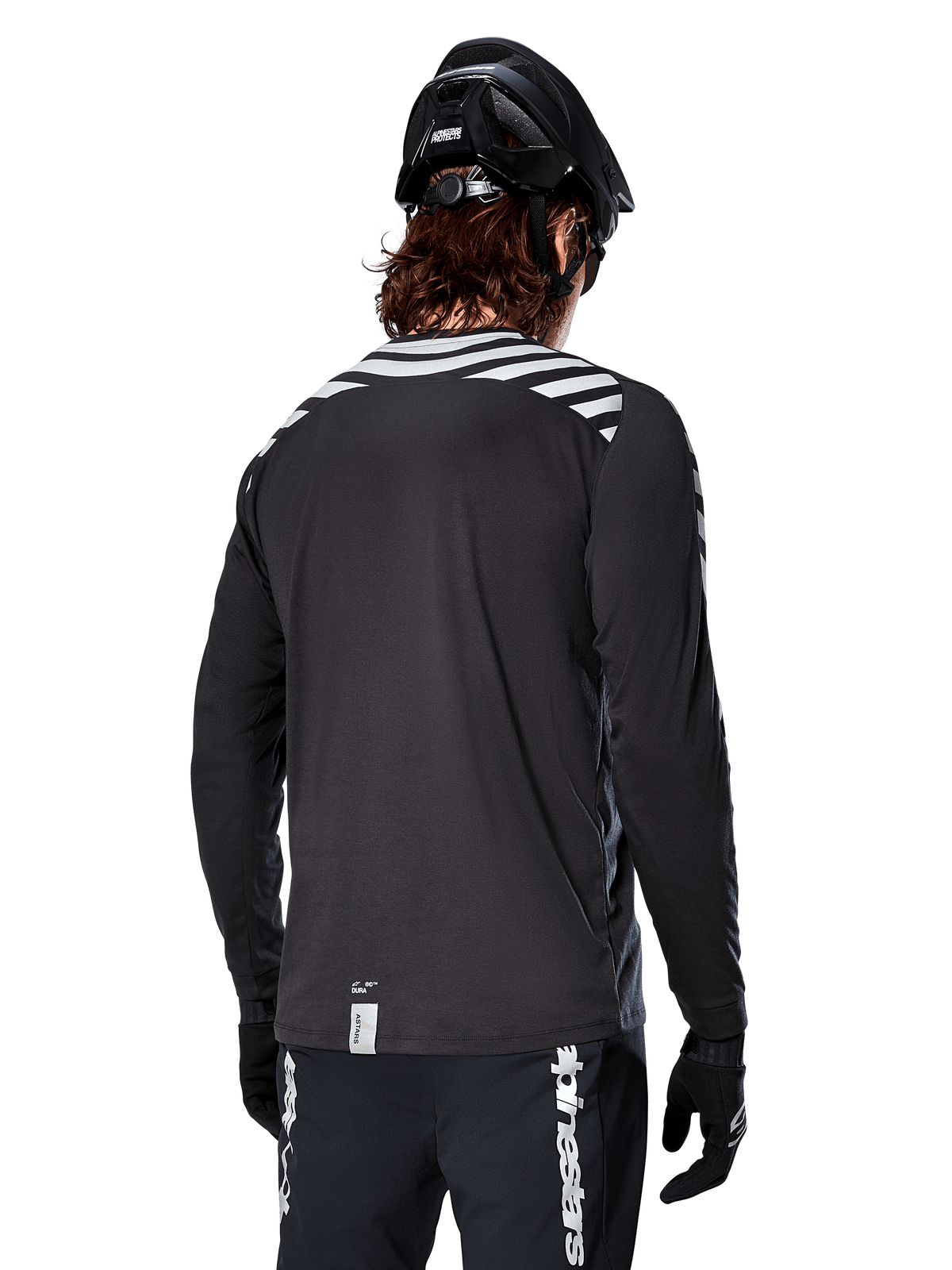 Camisola de Manga Comprida Alpinestars A-Dura Dri Oscar, preto refletor, vista traseira com riscas diagonais refletoras nos ombros e parte traseira mais comprida, usada com capacete e luvas de BTT pretos, equipamento técnico para BTT