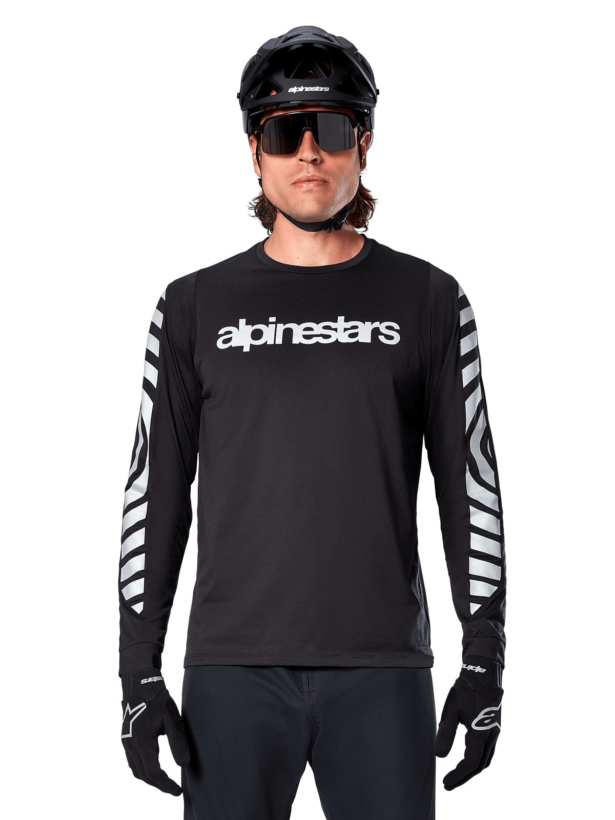 Ciclista masculino com Camisola Alpinestars A-Dura Dri Oscar de Manga Comprida, preto refletor, com logótipo Alpinestars branco no peito e gráficos prateados refletores nas mangas, acompanhado por capacete de BTT preto, óculos de sol e luvas pretas, equipamento de ciclismo de performance
