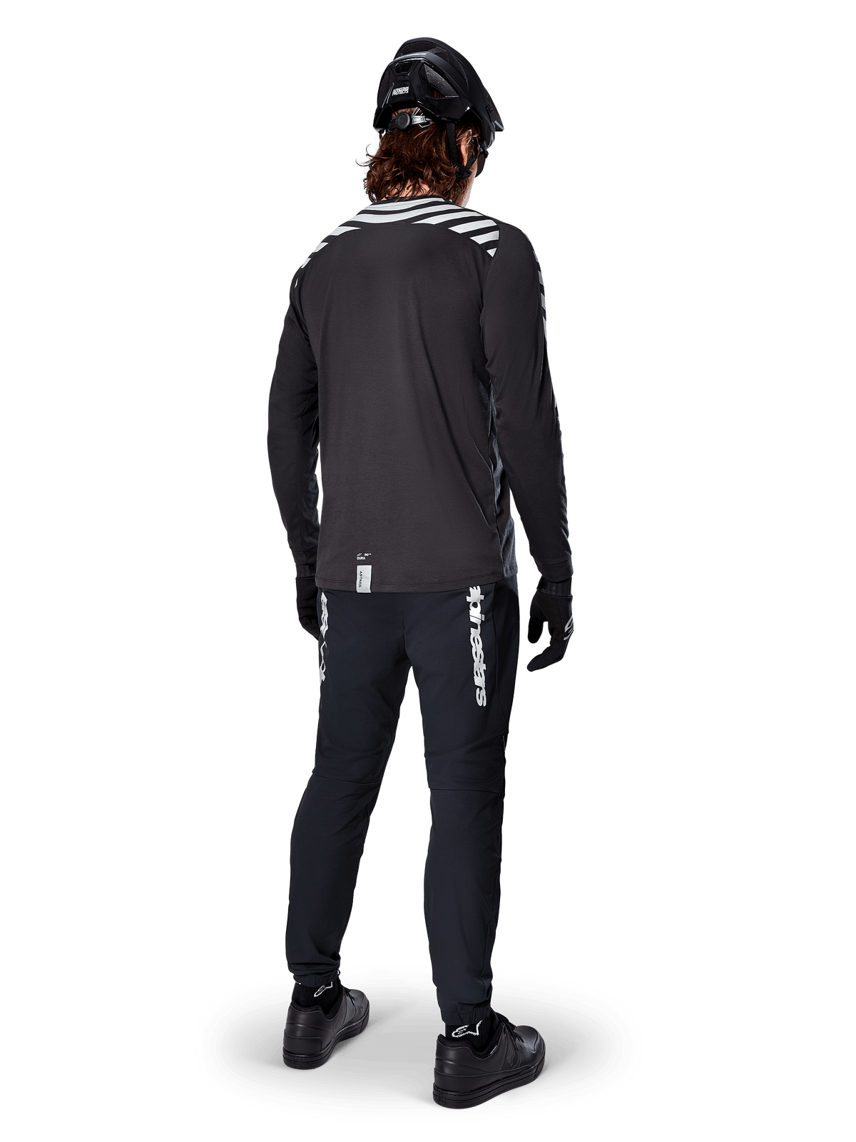Ciclista de BTT com a camisola de manga comprida Alpinestars A-Dura Dri Oscar em Preto Refletor, vista de costas, calças técnicas pretas, capacete e luvas, com riscas refletoras nos ombros e cauda mais longa para ciclismo todo-o-terreno.