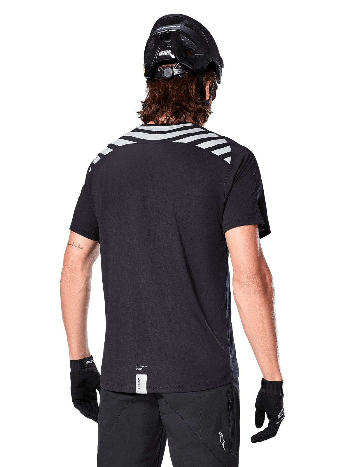 Alpinestars A-Dura Dri Oscar Jersey, camisola de MTB de manga curta, preto refletor, vista traseira num modelo a usar capacete de ciclismo e luvas, com riscas cinzentas refletoras nos ombros e cauda mais longa, tecido técnico com gestão de humidade para mountain bike