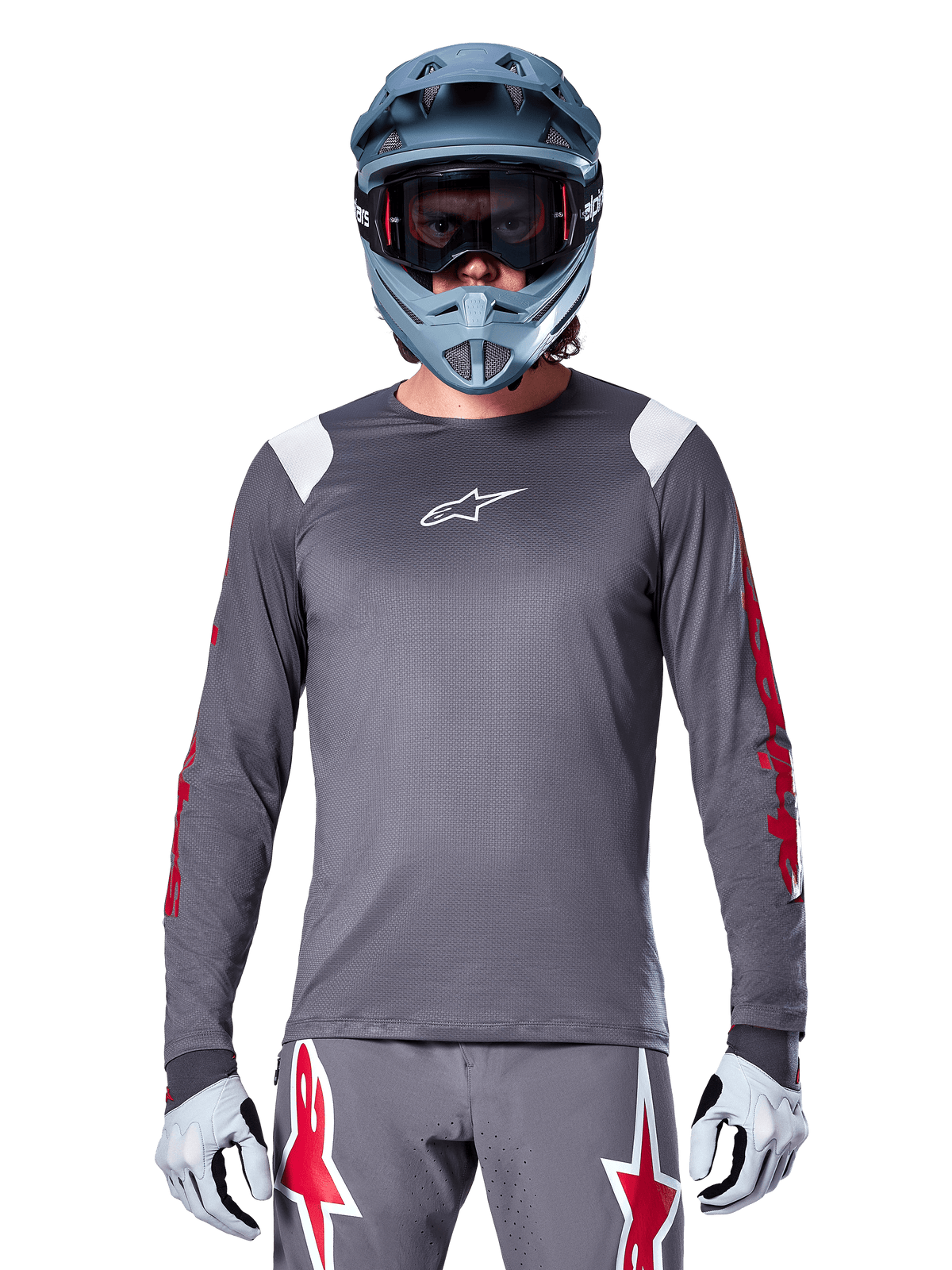 Ciclista de BTT a usar camisola de manga longa Alpinestars A-Supra Astar, cinzento escuro com grafismos vermelhos nas mangas e detalhes brancos nos ombros, calças cinzentas a condizer com logótipos de estrela vermelhos, capacete integral azul e óculos de proteção, equipamento de ciclismo todo-o-terreno coordenado