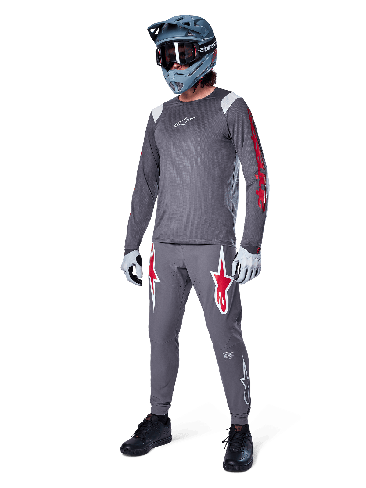 Camisola de Manga Comprida Alpinestars A-Supra Astar em cinzento escuro, com branding vermelho nas mangas e logótipo branco no peito, usada por um ciclista com calças cinzentas a condizer, capacete integral cinza-azulado e luvas de performance, equipamento coordenado de BTT para gravity