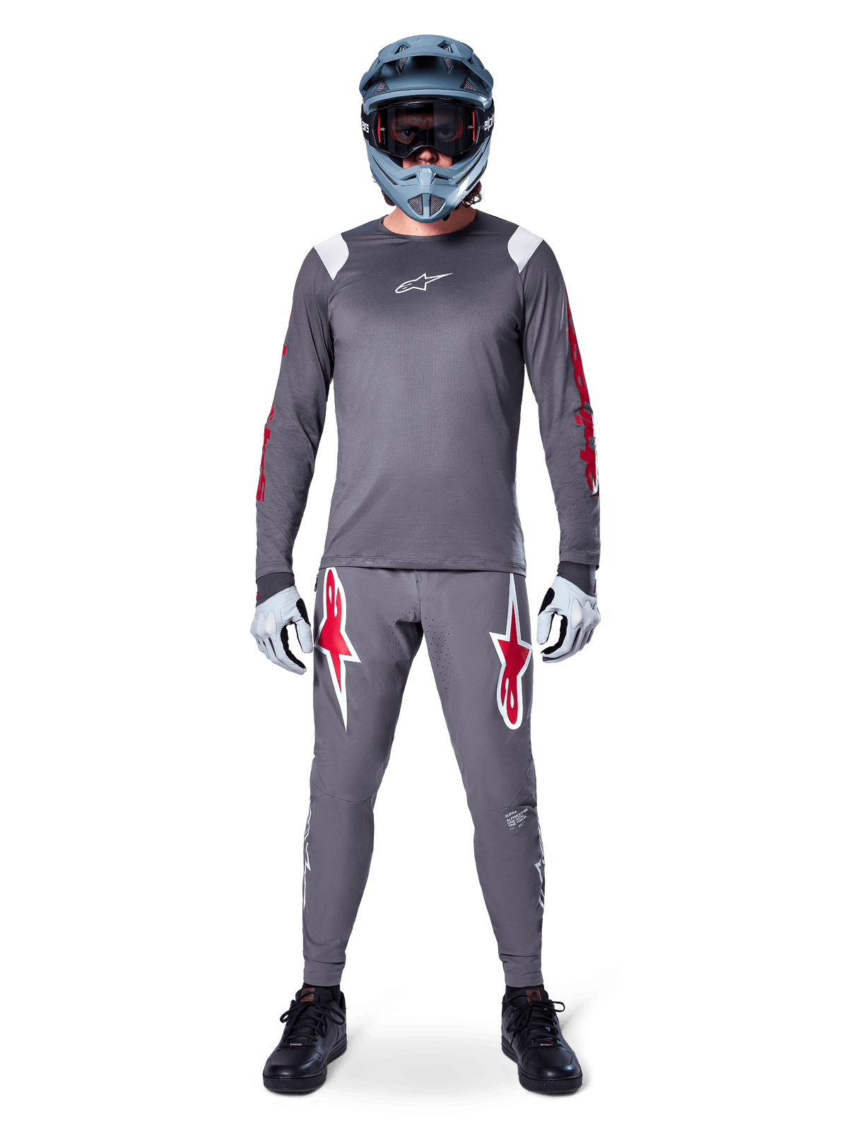 Ciclista de MTB com camisola de manga comprida Alpinestars A-Supra Astar, cinzento escuro com pormenores vermelhos e logótipo branco no peito, calças cinzentas a condizer com grandes grafismos de estrelas vermelhas, capacete integral azul com óculos, luvas cinza claro e sapatos pretos, conjunto de equipamento profissional de mountain bike