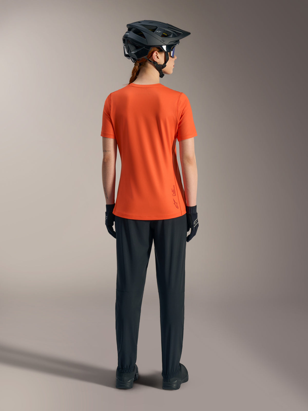 Alpinestars Women's Stella A-Dura Ride Short Sleeve Jersey in orange, back view with black MTB pants and helmet.