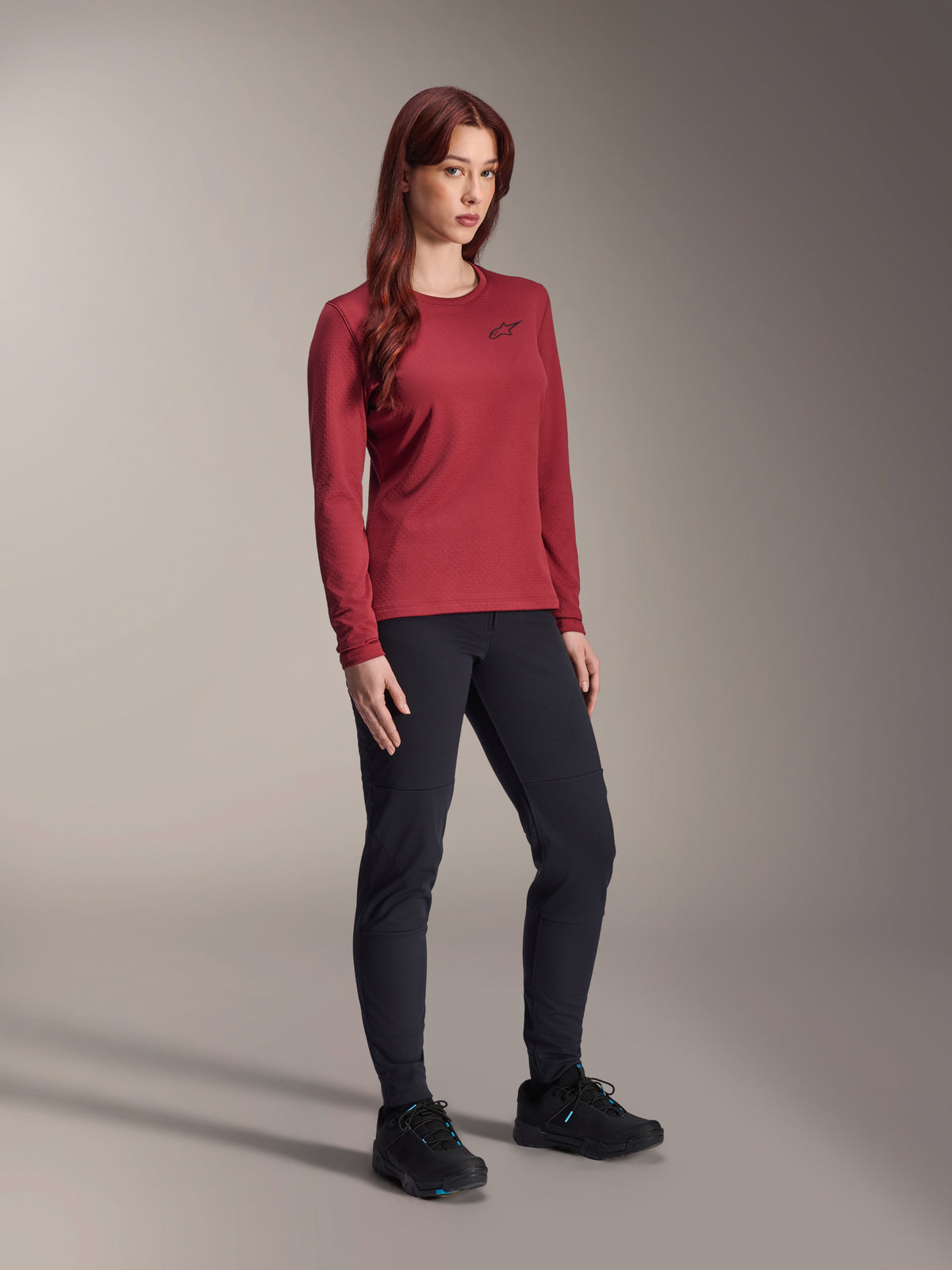 Mulher a usar calças térmicas Alpinestars Stella A-Dura em preto e camisola térmica Stella A-Dura de manga comprida em bordeaux, vestuário técnico de BTT para condução em climas frios, design de corte justo em material durável, fundo de estúdio