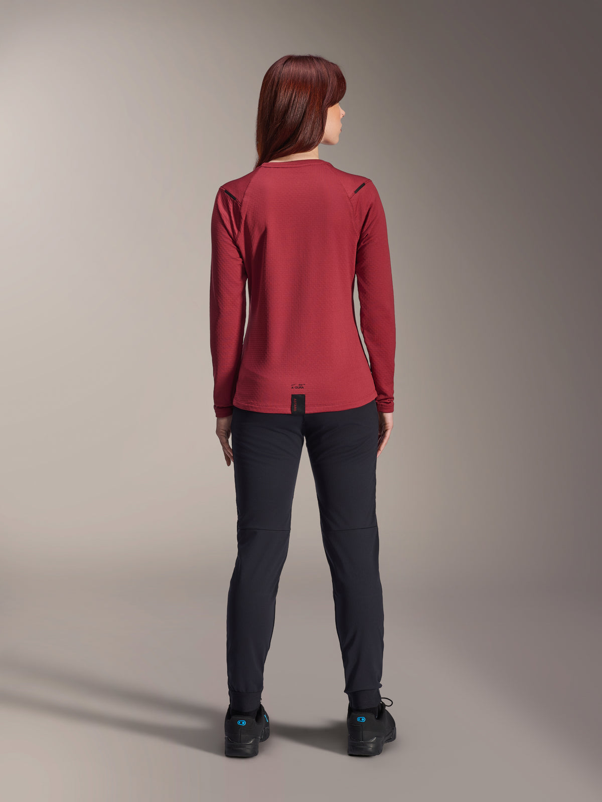 Mulher a usar a camisola térmica Alpinestars Stella A-Dura, Borgonha, vista traseira, camisola de ciclismo de manga comprida com isolamento e cauda alongada, combinada com calças pretas e sapatos de ciclismo pretos, vestuário de BTT para exterior.