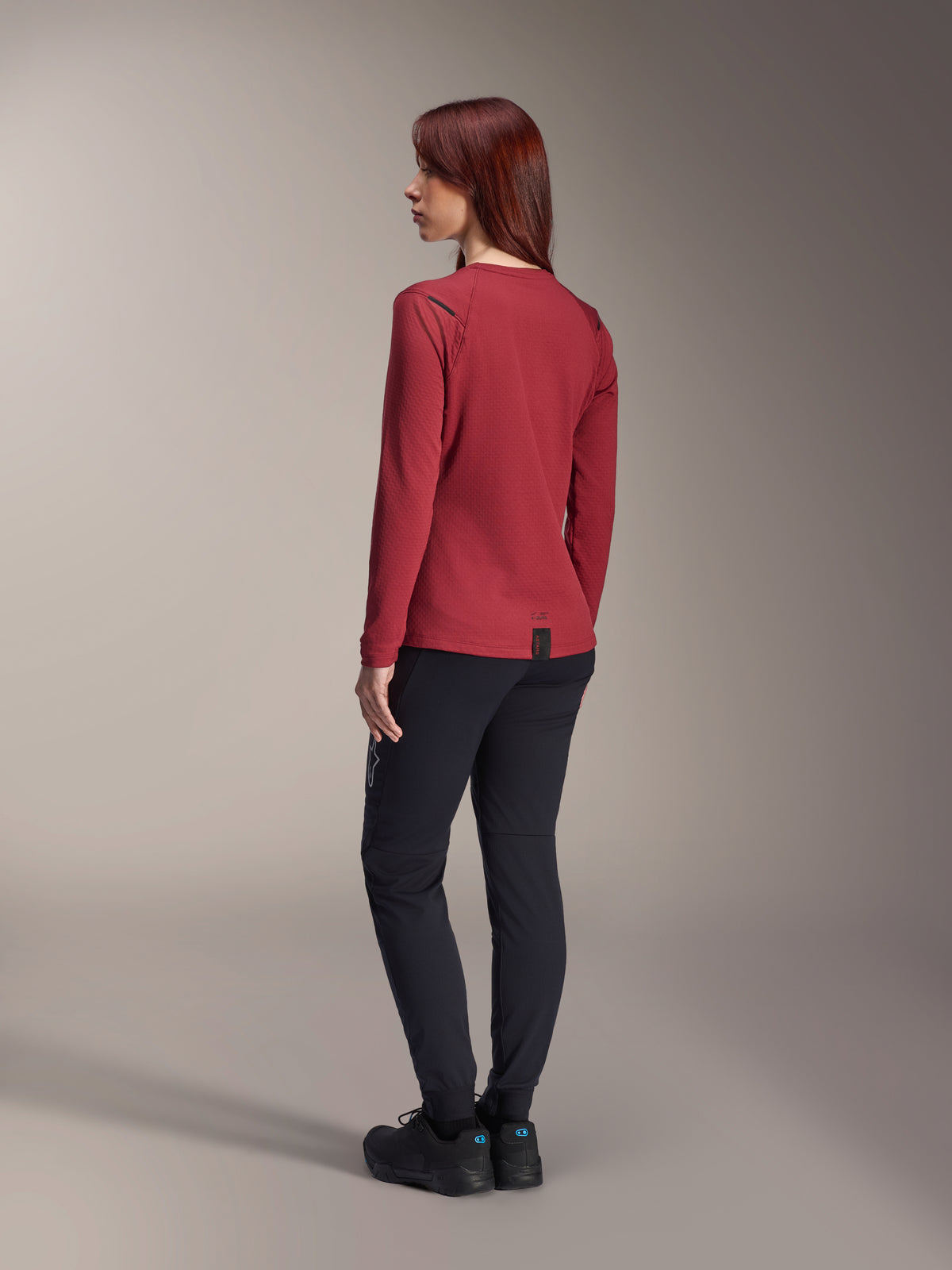 Mulher a usar Calças Térmicas Alpinestars Stella A-Dura em preto, combinadas com uma camisola térmica de manga comprida vermelha e sapatos pretos, vista traseira, vestuário de BTT para condução em clima frio.